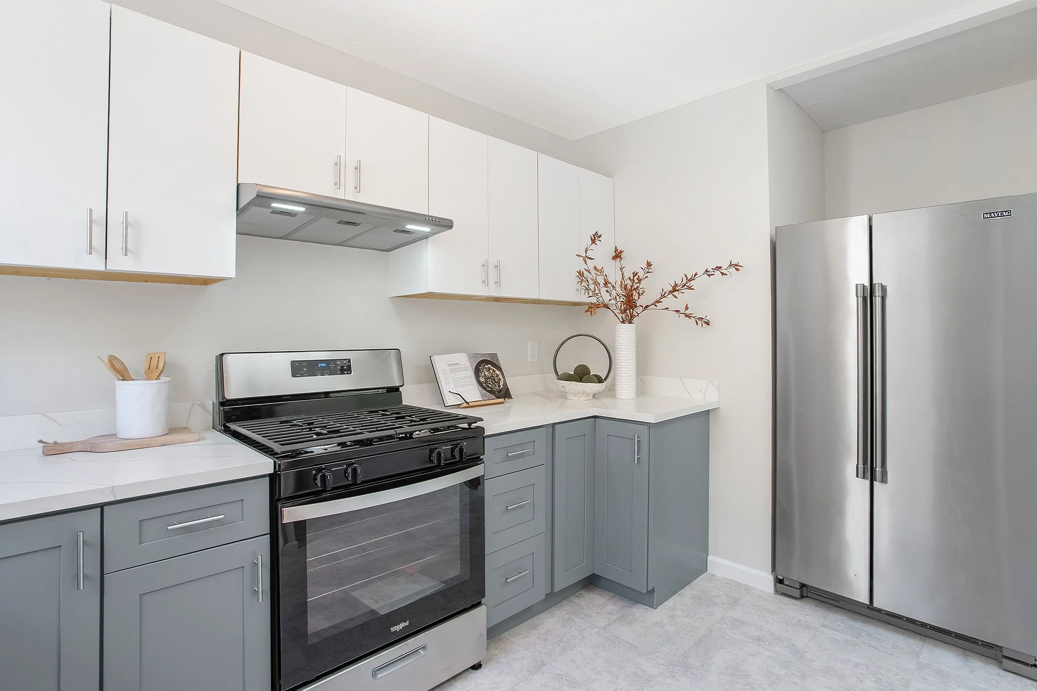 Modern kitchen with white upper cabinets, gray lower cabinets, stainless steel refrigerator, black stove, and decorative accents including a vase with leaves and a bowl of green balls.