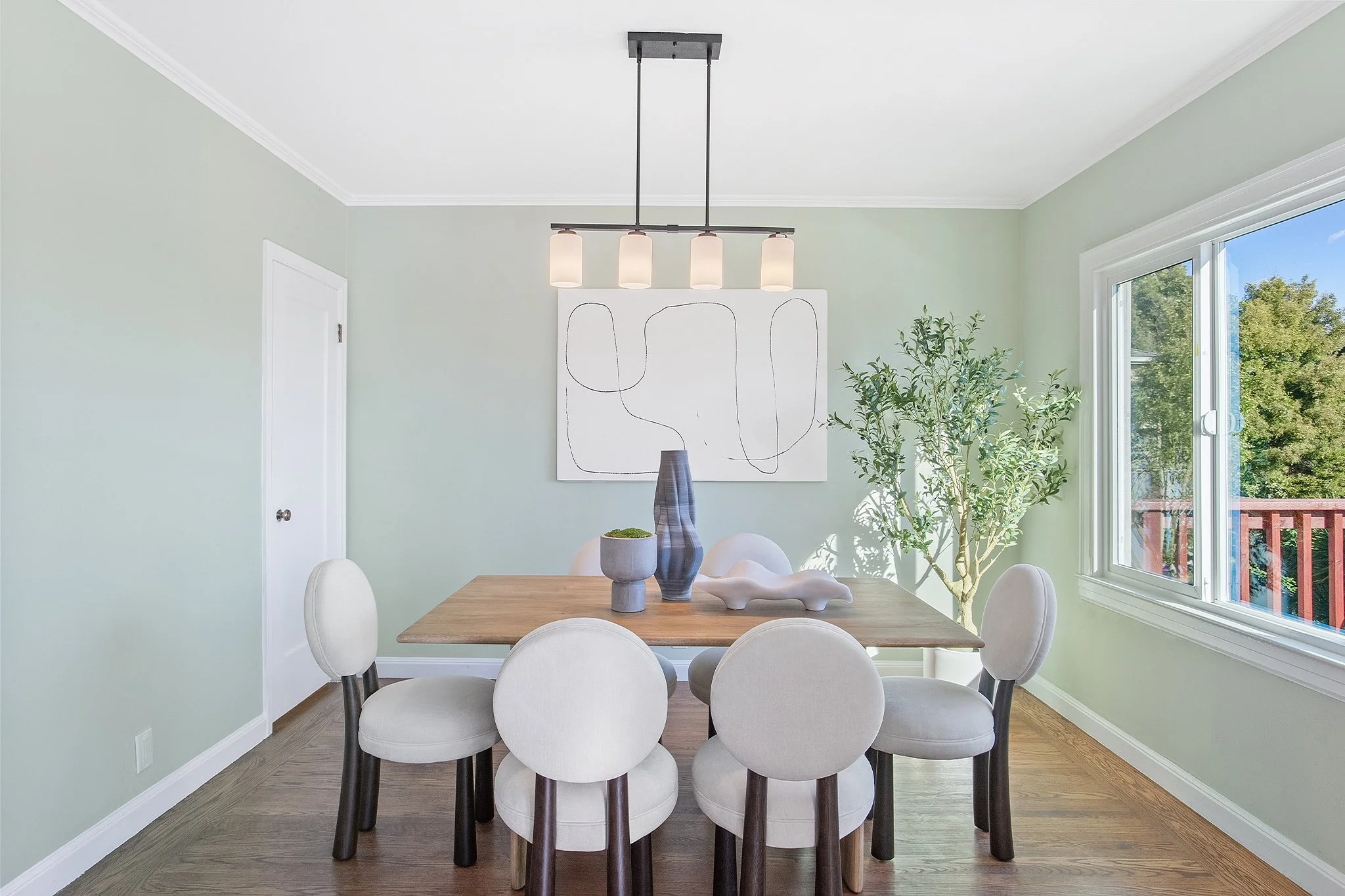 Dining room with a wooden table, six white upholstered chairs, modern light fixture, abstract art on the wall, potted plant, and a large window showing greenery outside.
