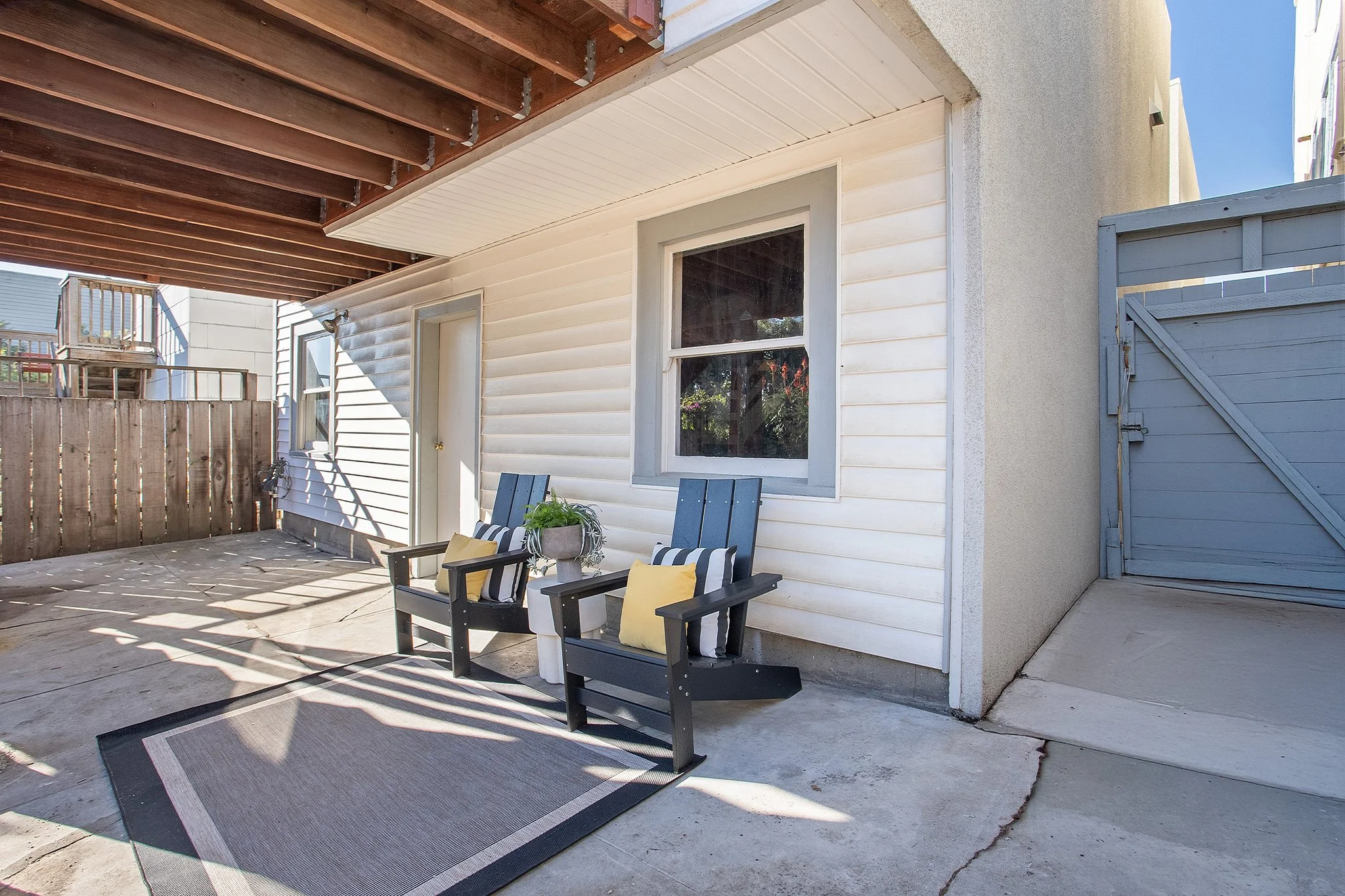 Back porch with two black chairs with yellow and black and white pillows, a small table with a potted plant, and a gray rug, attached to a white house with a window and door, wooden fence, and blue gate, under a wooden balcony overhang.