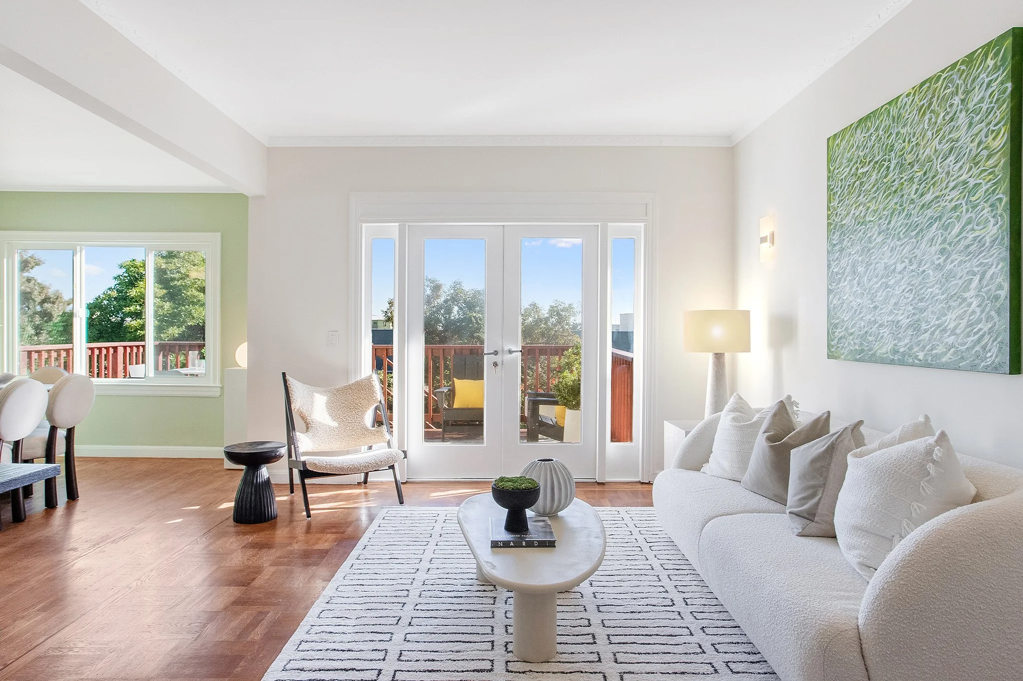 Bright living room with white walls, wooden floor, glass double doors leading to a balcony with outdoor seating, and large windows. Furnished with a white sofa, a round coffee table with decorative vases, a standing lamp, and a wall-mounted artwork. 