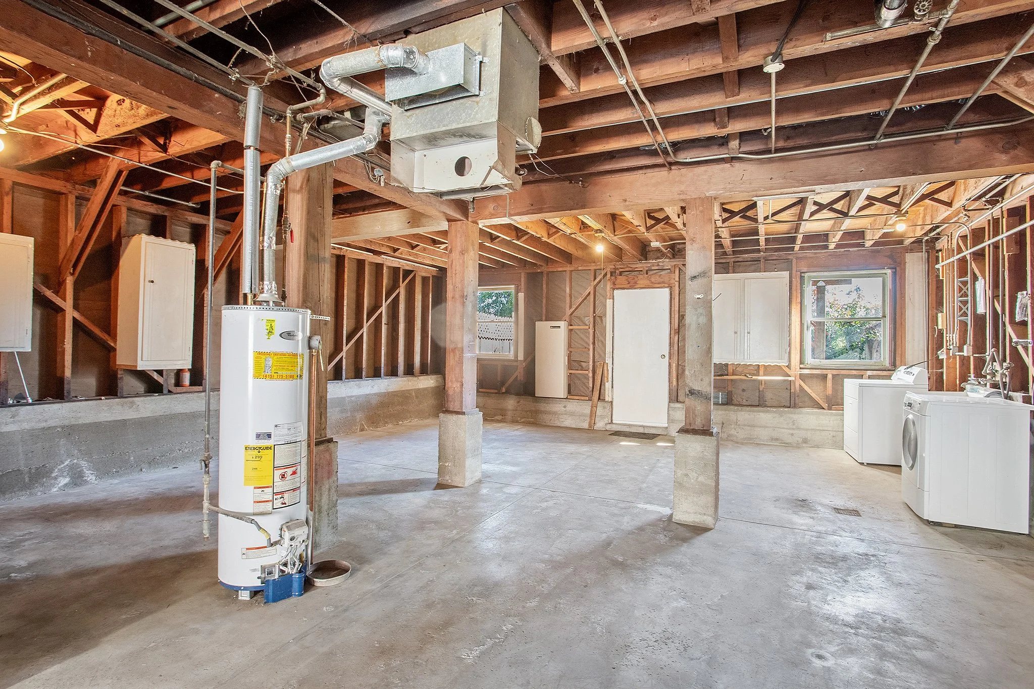 Empty unfinished basement with exposed wooden beams, concrete floor, water heater, laundry area with washing machine, and a window.