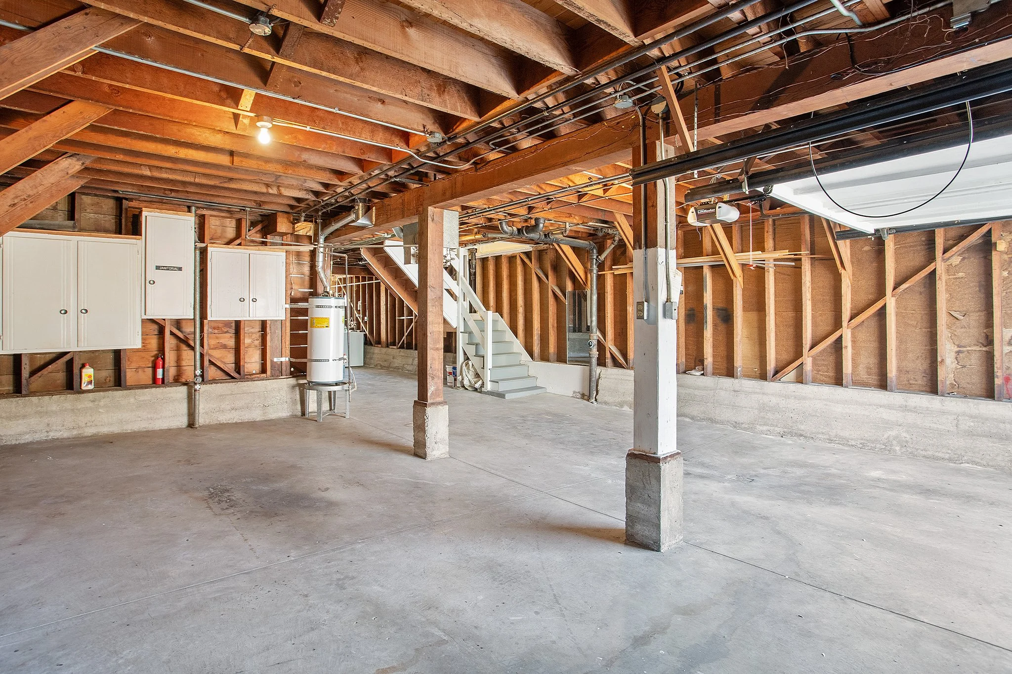 Empty unfinished garage with exposed wooden ceiling beams, concrete floor, utility panels, water heater, and stairway.