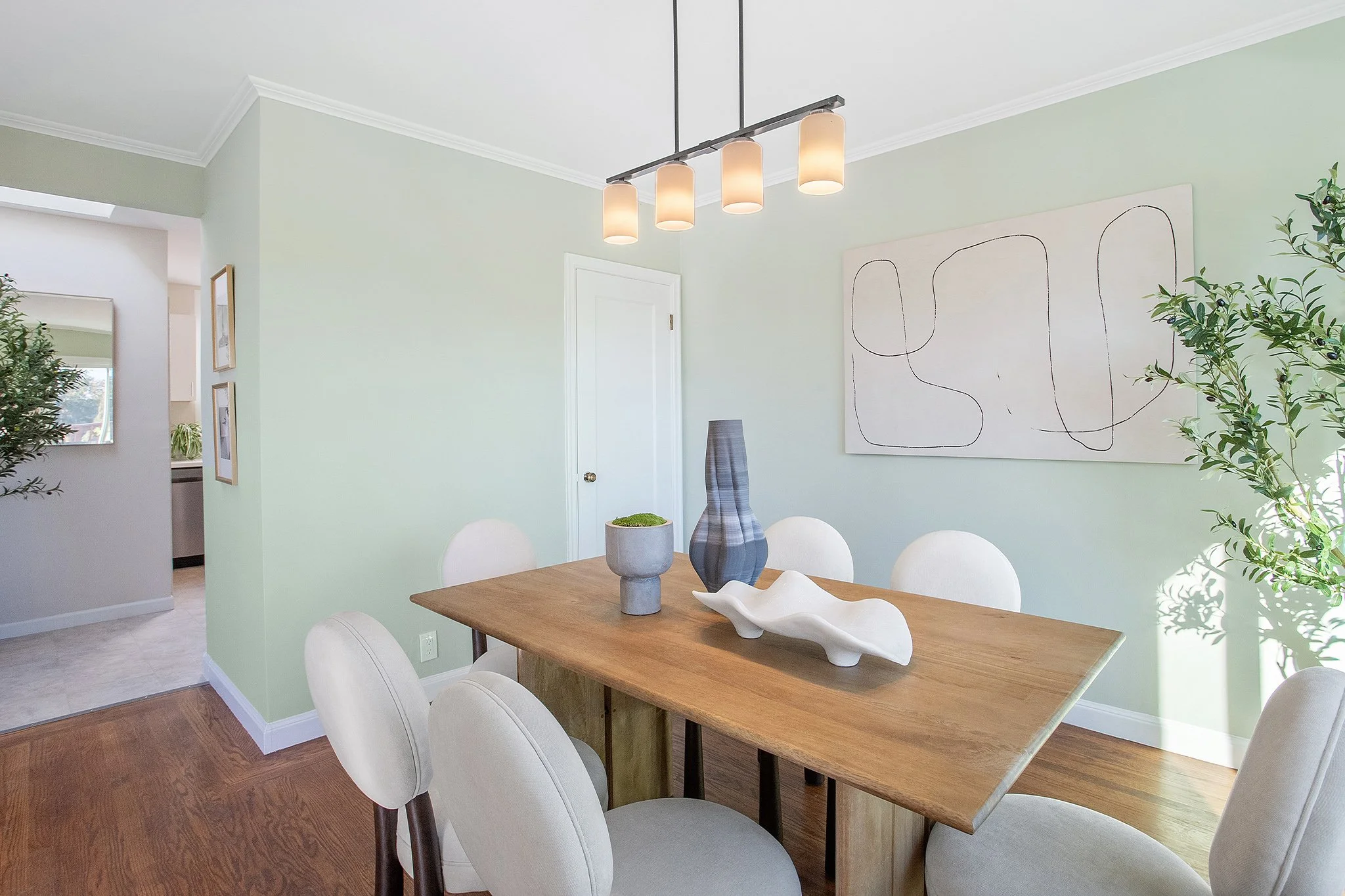Dining room with a wooden table, six cream-colored chairs, modern light fixture, abstract artwork on mint green wall, and decorative vases and sculpture on the table.