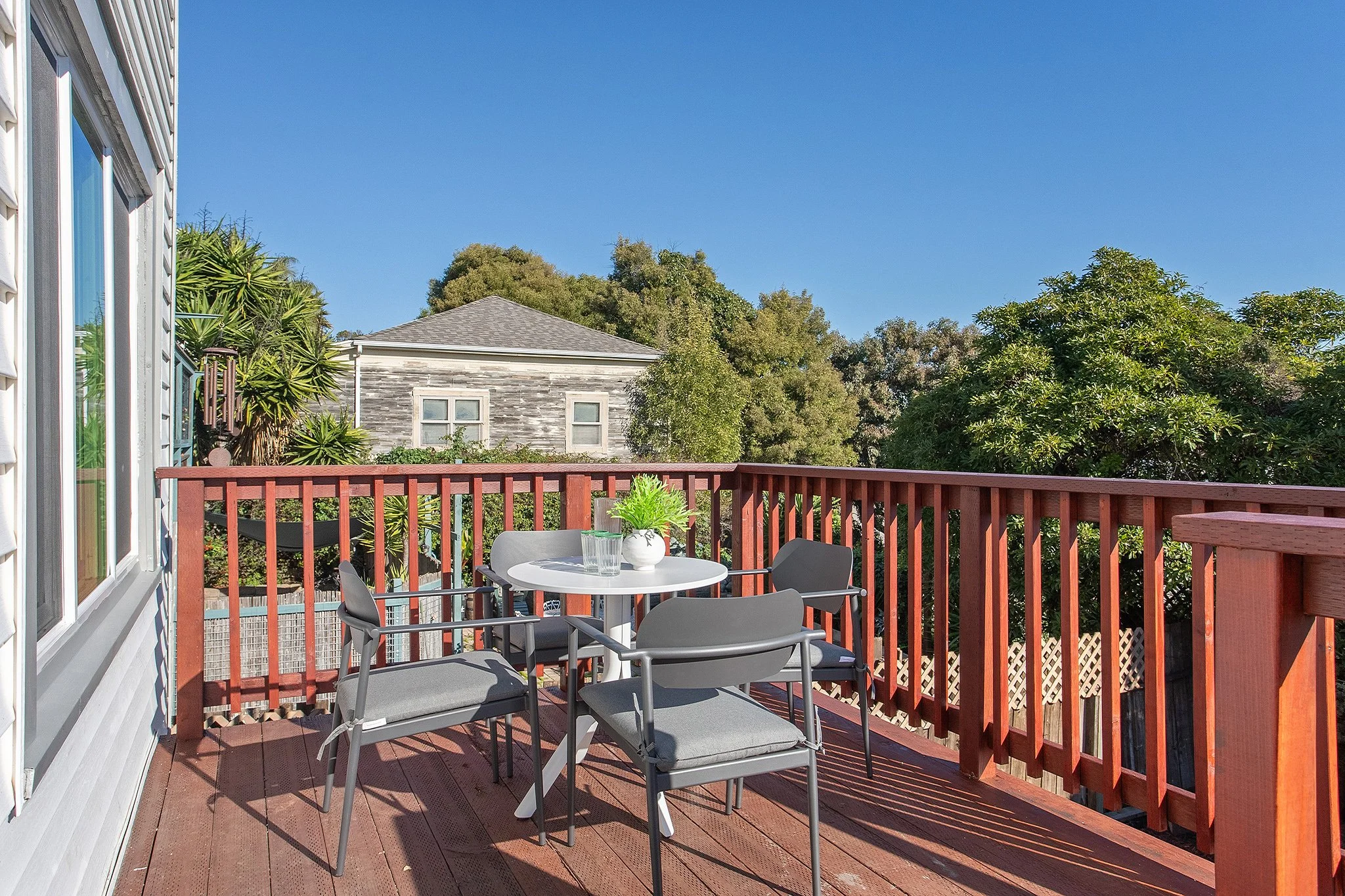 A wooden balcony with a round table, four chairs, a potted plant, and a glass of water, overlooking trees and neighboring houses under a clear blue sky.