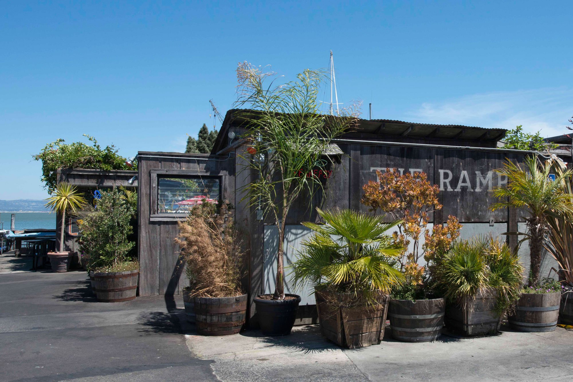 A rustic wooden building with outdoor seating, surrounded by potted plants and trees, with a view of water and boats in the background under a clear blue sky.