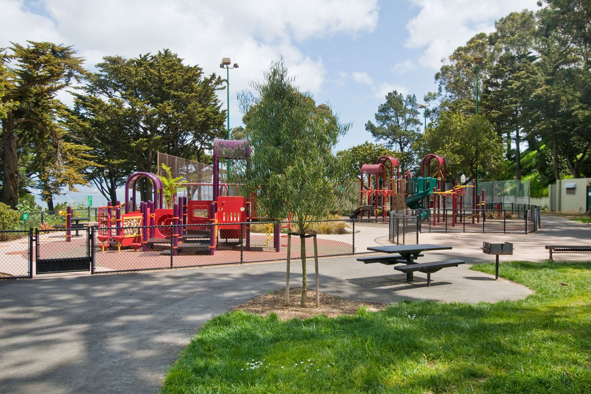 Colorful playground with slides and climbing structures, surrounded by trees and fencing, in a park setting on a sunny day.