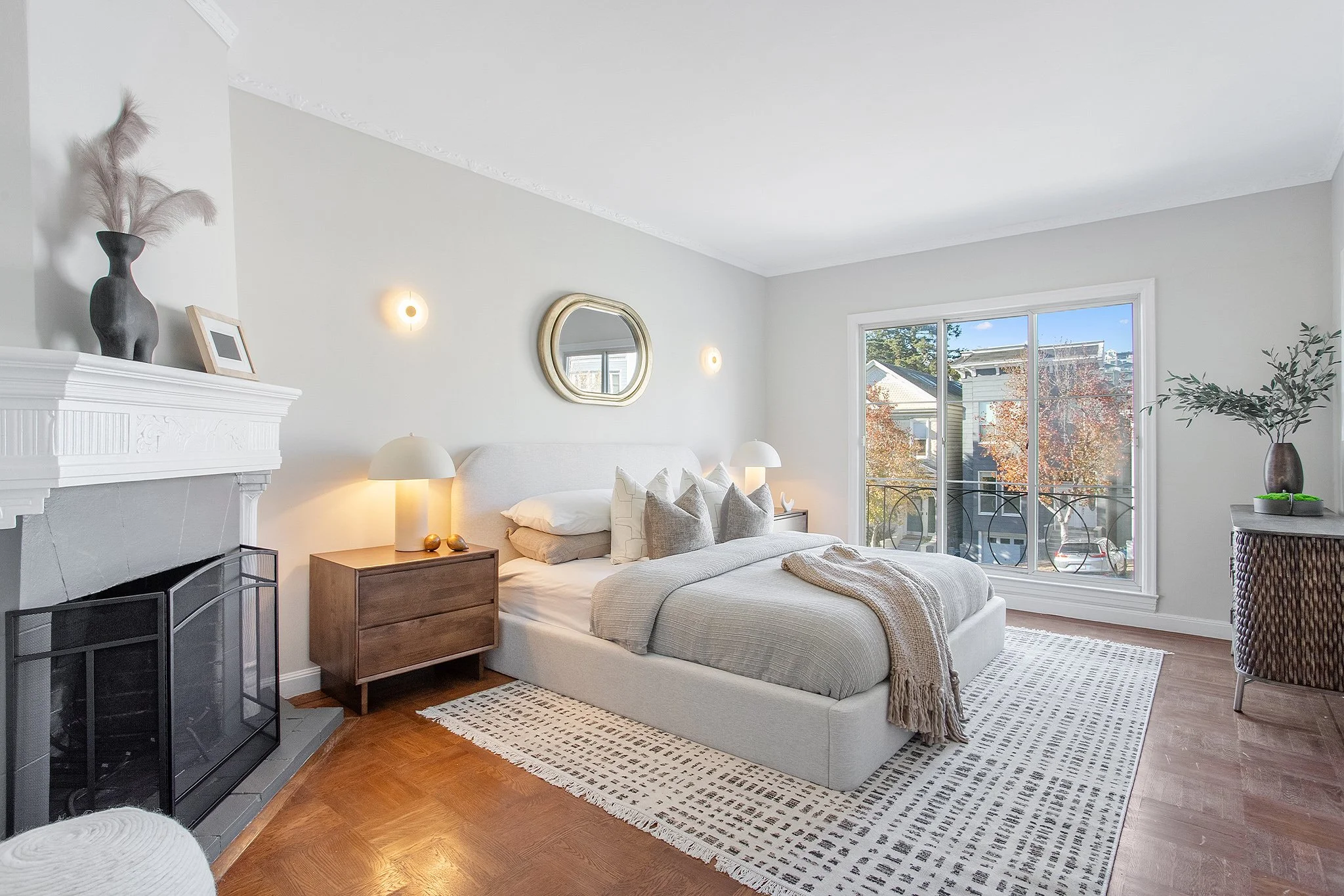 Bright bedroom with large window, white walls, bed with beige and gray bedding, two wooden nightstands with lamps, decorative mirror, fireplace, potted plant, and modern decor.