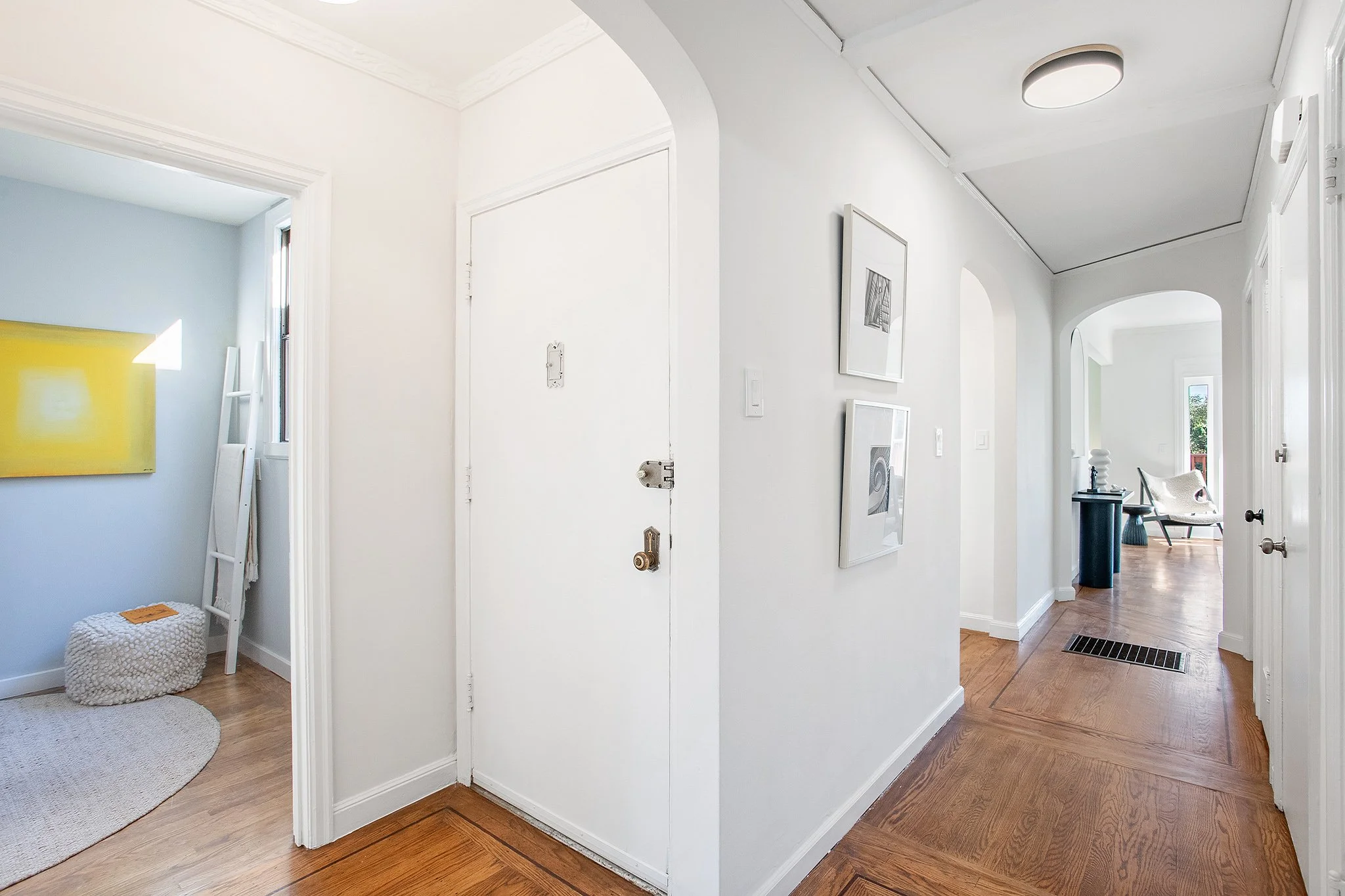 Entrance hallway of a home with hardwood floors, white walls, and framed artwork; leads to a sitting area with a chair and side table in the background.