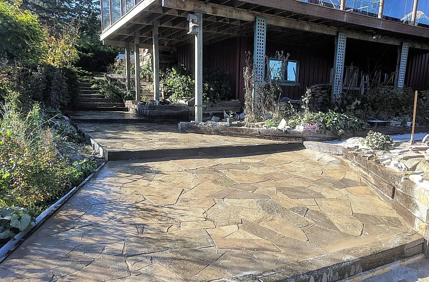 Outdoor patio with stone flooring, garden beds, and a house with a deck in the background.