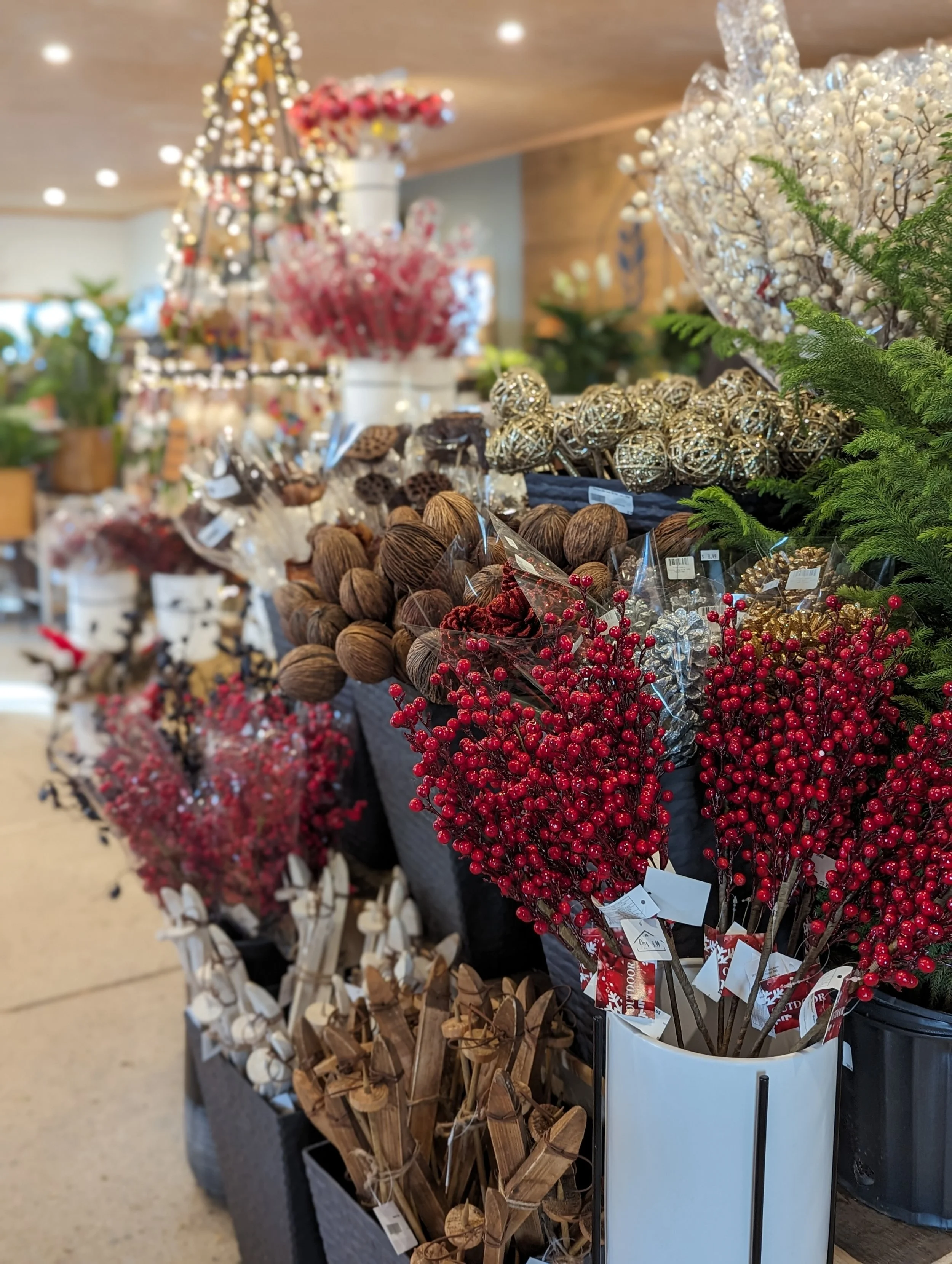 Display of various holiday floral and decorative arrangements including red berries, brown nuts, white branches, and wrapped wooden sticks, with a Christmas tree in the blurred background.