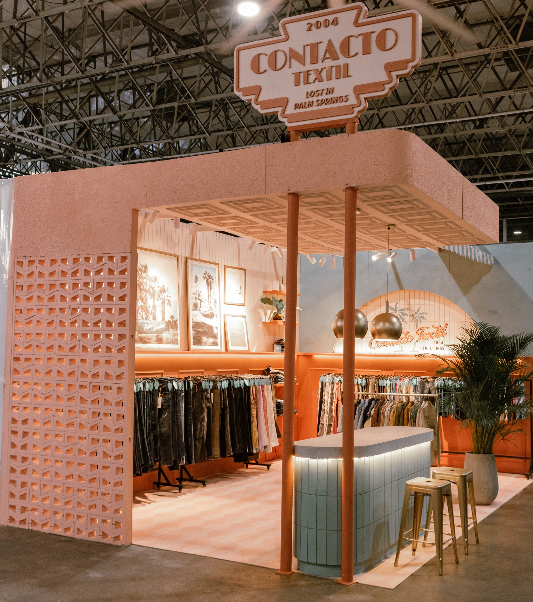 Stand de ropa en un evento, con un letrero que dice "2004 Contacto Textil, Perdido en Palm Springs" y decoraciones en tonos pastel, con cuadros en la pared y plantas decorativas.