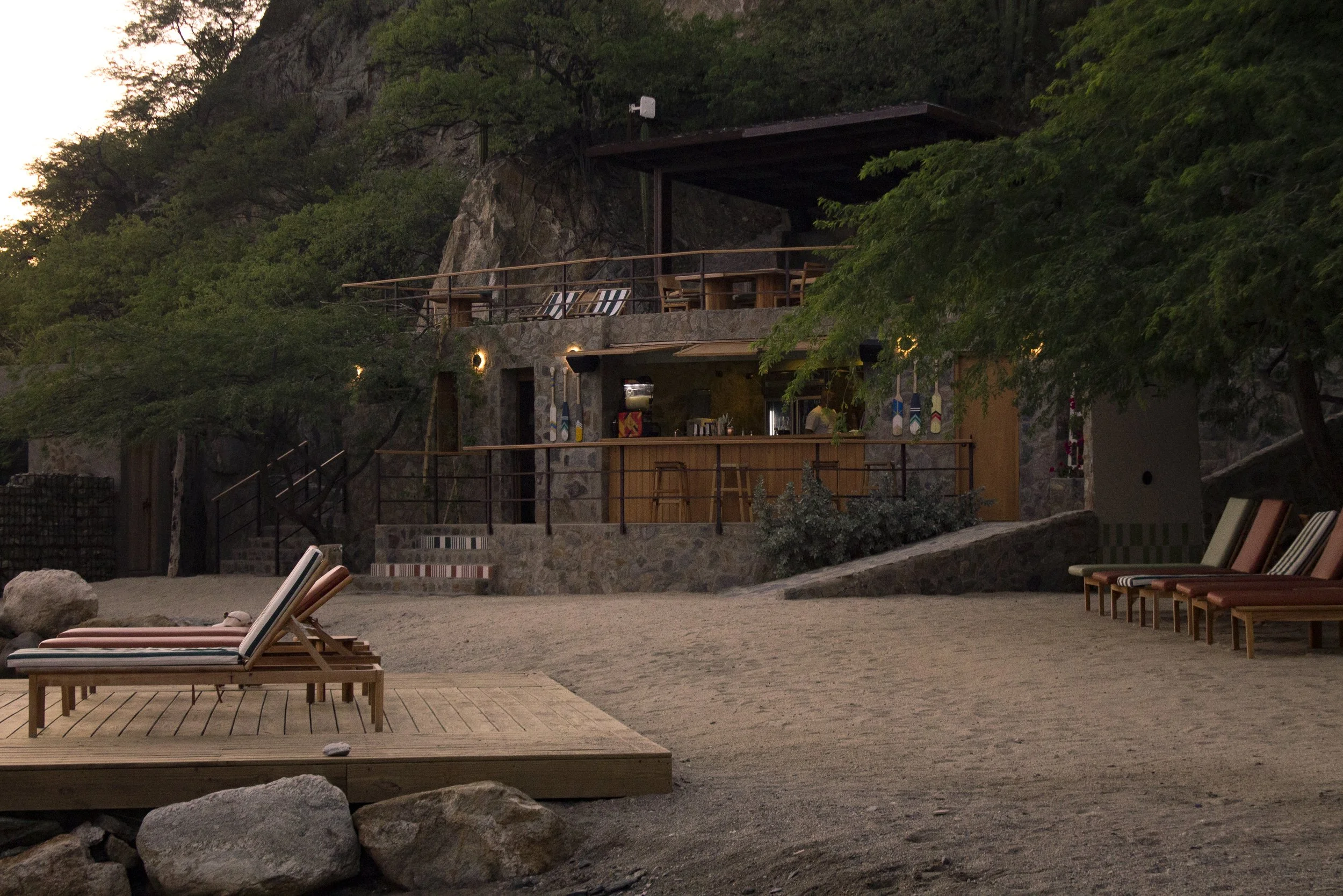 Una vista de un establecimiento de piedra y madera en una ladera con árboles alrededor, con un área de descanso con sillas y camastros de madera en la playa, durante el atardecer