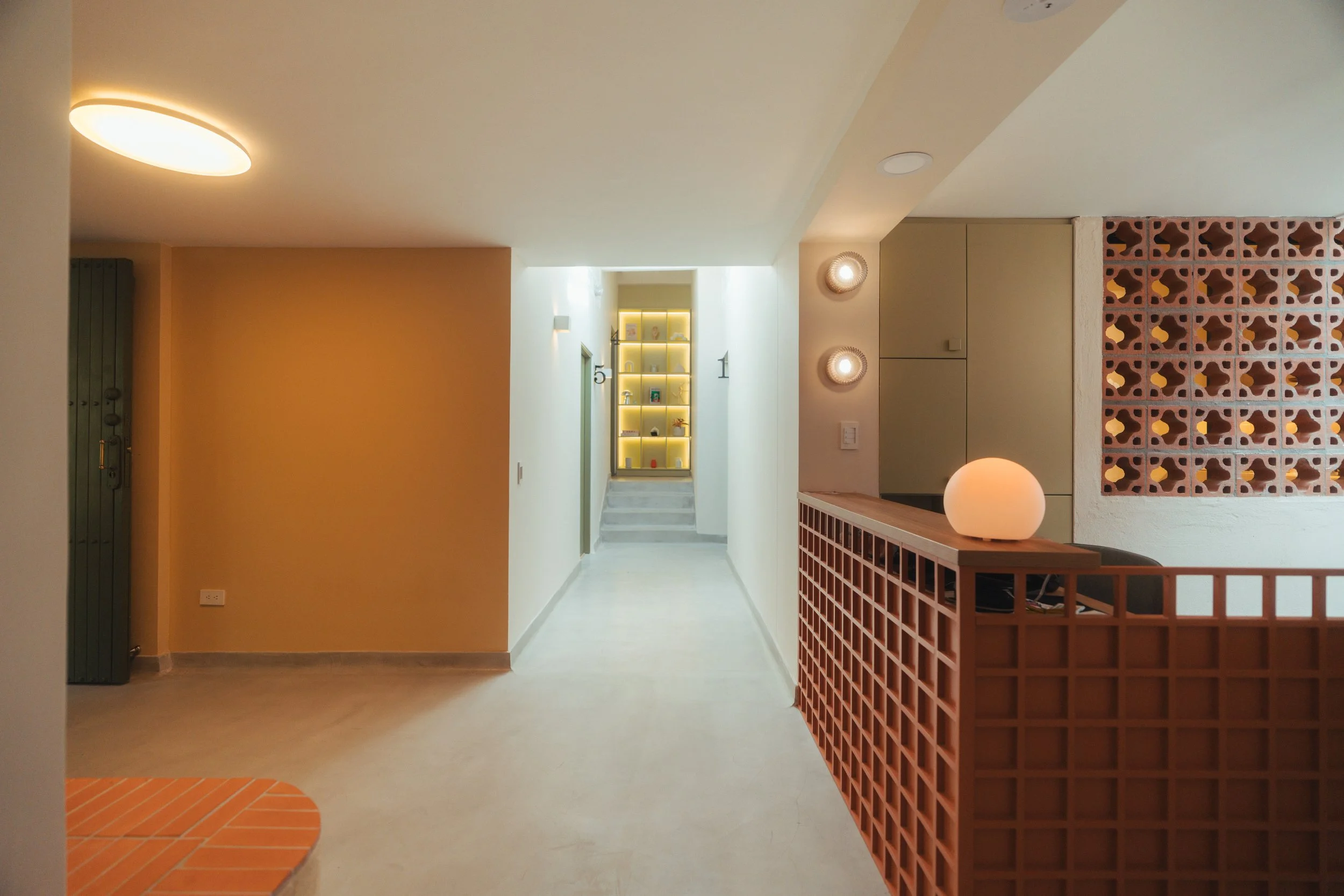 Interior de un vestíbulo moderno con pared amarilla, escalera con estantería iluminada, paredes blancas, lámparas circulares y un mostrador con diseño geométrico.