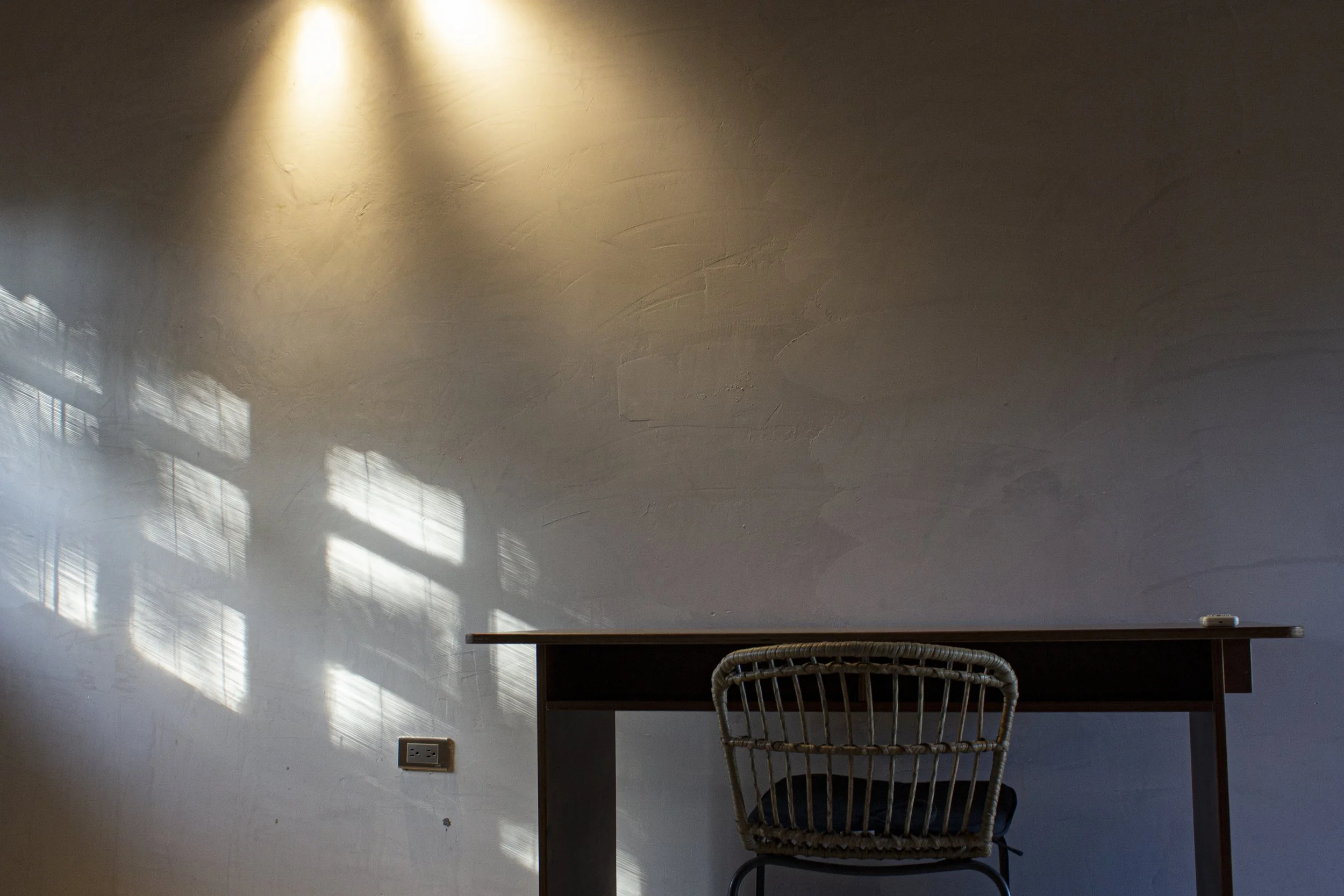 Una pared de color claro con sombras de ventanas y una lámpara en el techo, una mesa de color oscuro con un objeto pequeño en la esquina, y una silla de mimbre frente a la mesa.