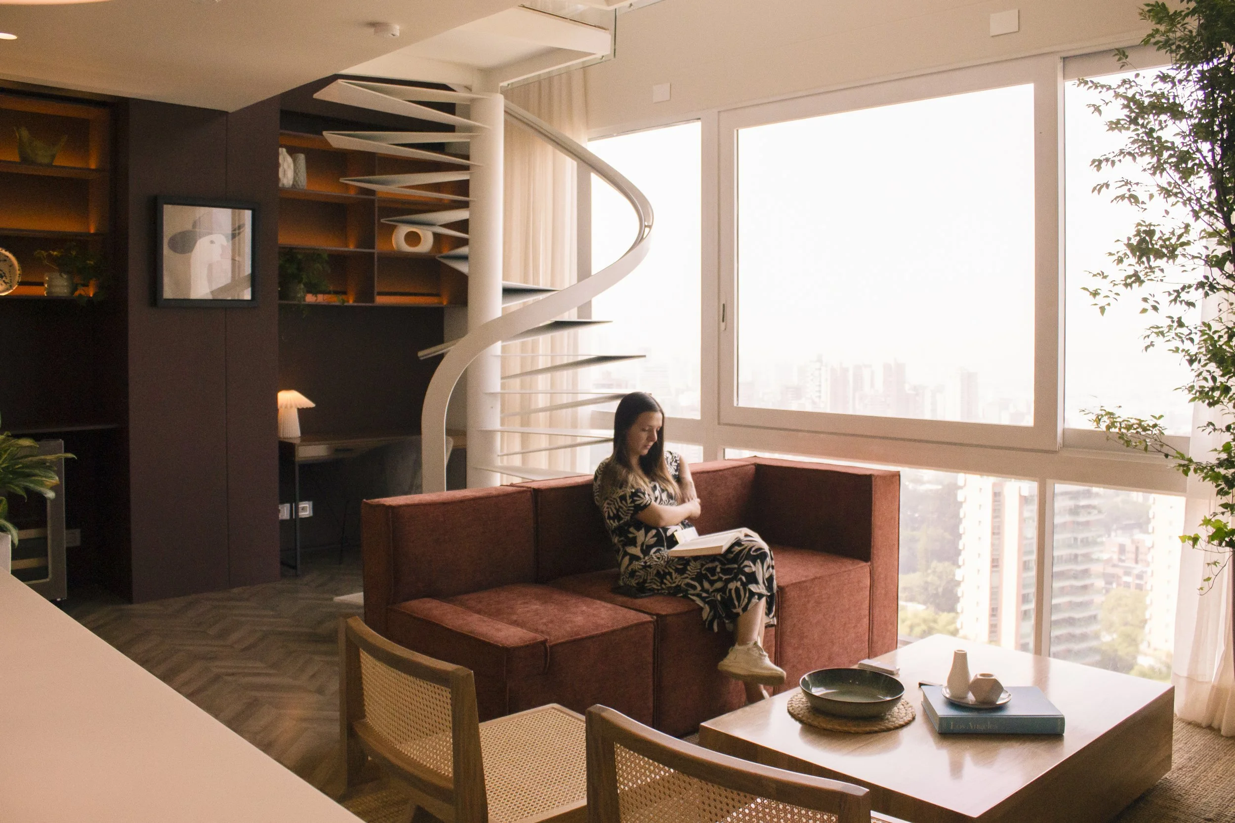 Una mujer sentada en un sofá de color marrón leyendo un libro en un apartamento con vista a la ciudad, grandes ventanas, un árbol y decoración moderna.