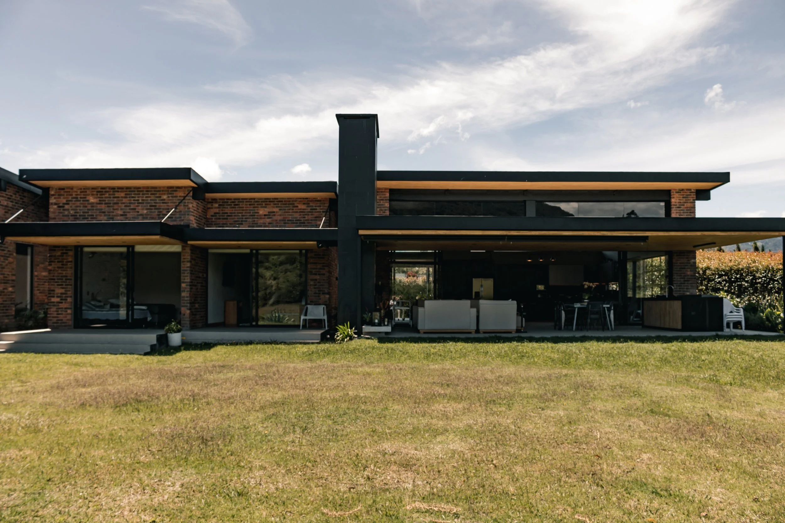 Casa moderna de dos pisos con muro de ladrillo y ventanas grandes, ubicada en un amplio jardín con césped, bajo un cielo parcialmente nublado.