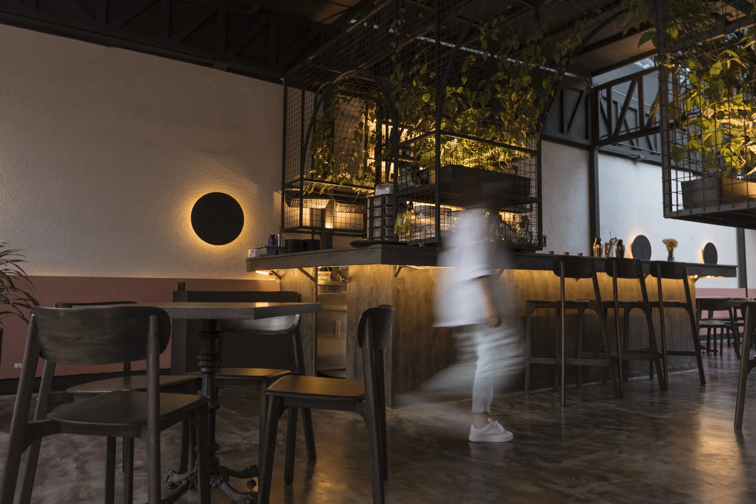 Interior de un restaurante moderno con iluminación cálida, mesas de madera y un bar de barandilla de madera y metal, con un camarero en movimiento borroso.