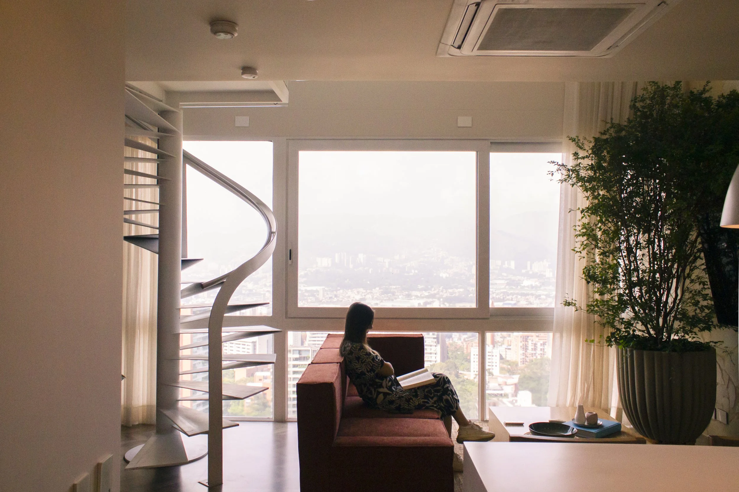 Una mujer sentada en un sofá leyendo un libro frente a una gran ventana con vista a la ciudad y las montañas en el fondo, en un apartamento moderno con plantas y decoración