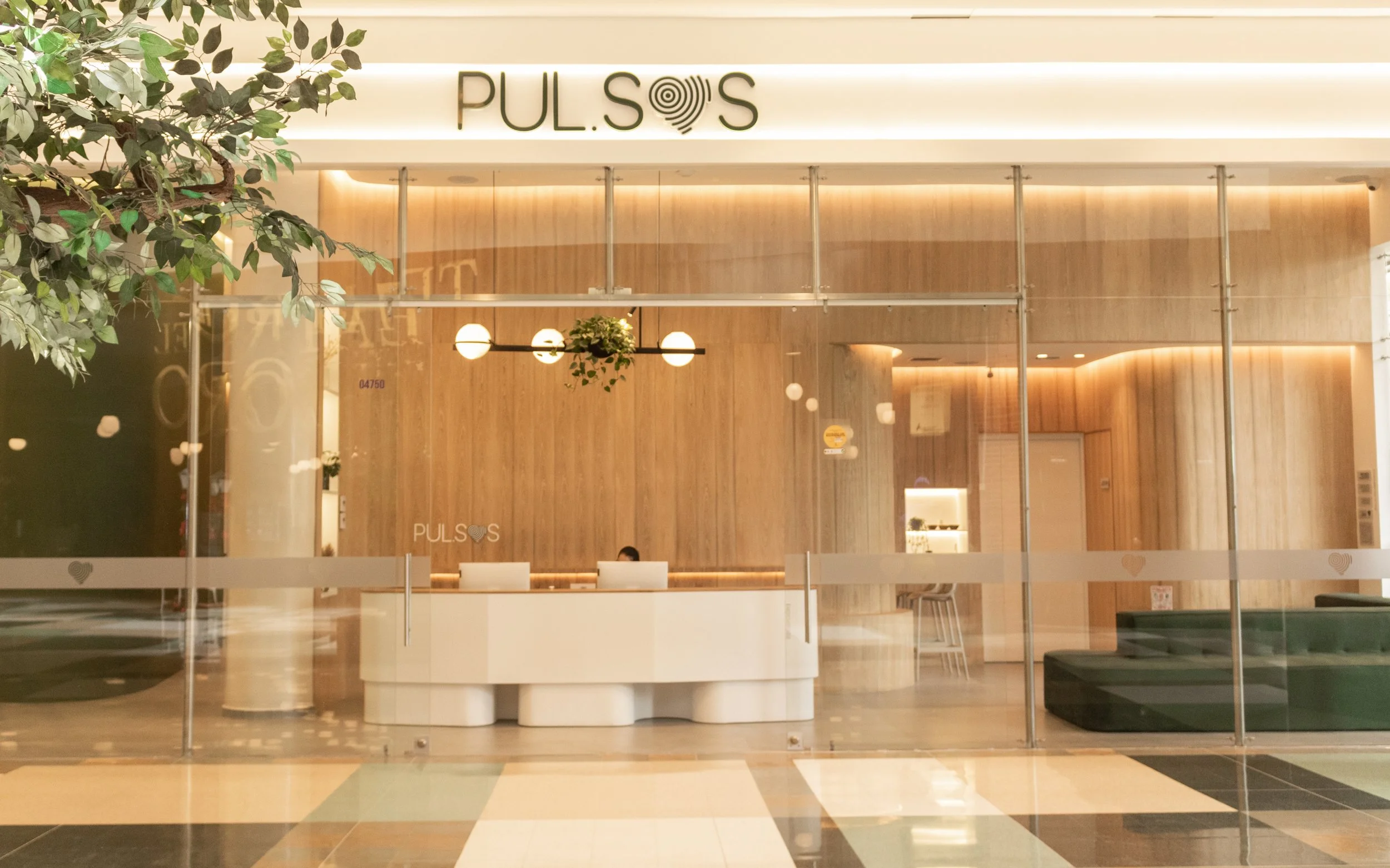 Interior de una sala de espera moderna con paredes de madera, mueble de recepción blanco y sillones verdes outdoor, decorada con iluminación cálida y plantas verdes, vista desde afuera a través de una estructura de vidrio.