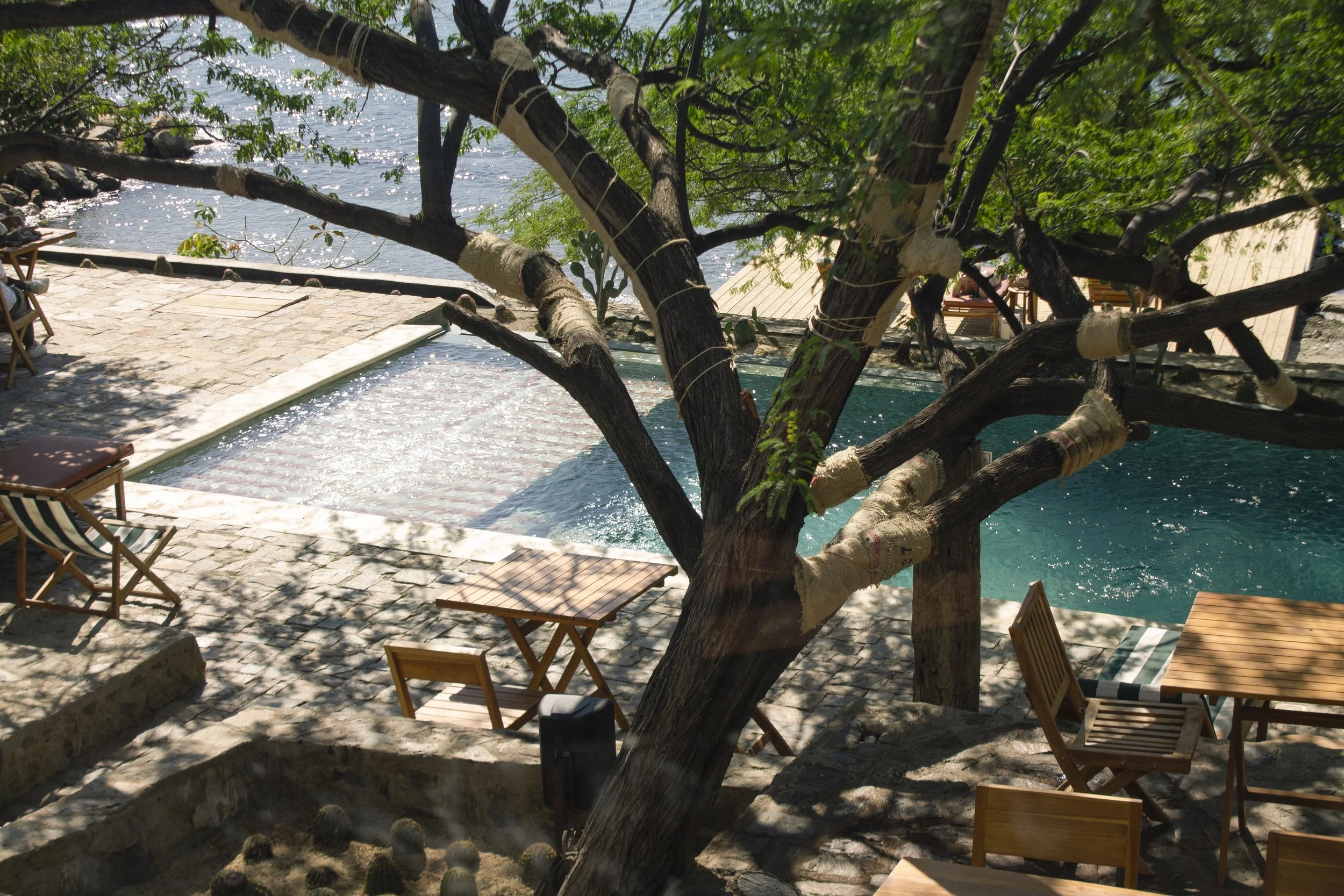 Vista de una piscina junto al río con sombra de un árbol y sillas de madera.