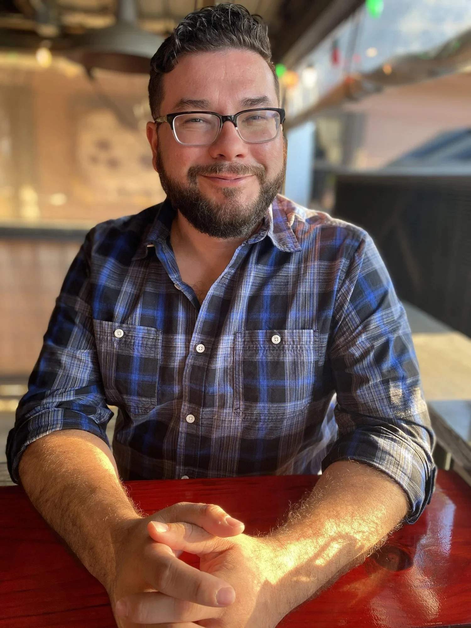 A man with glasses and a beard, wearing a plaid shirt, sitting at a table with hands clasped, smiling at the camera, in a warmly lit indoor setting.