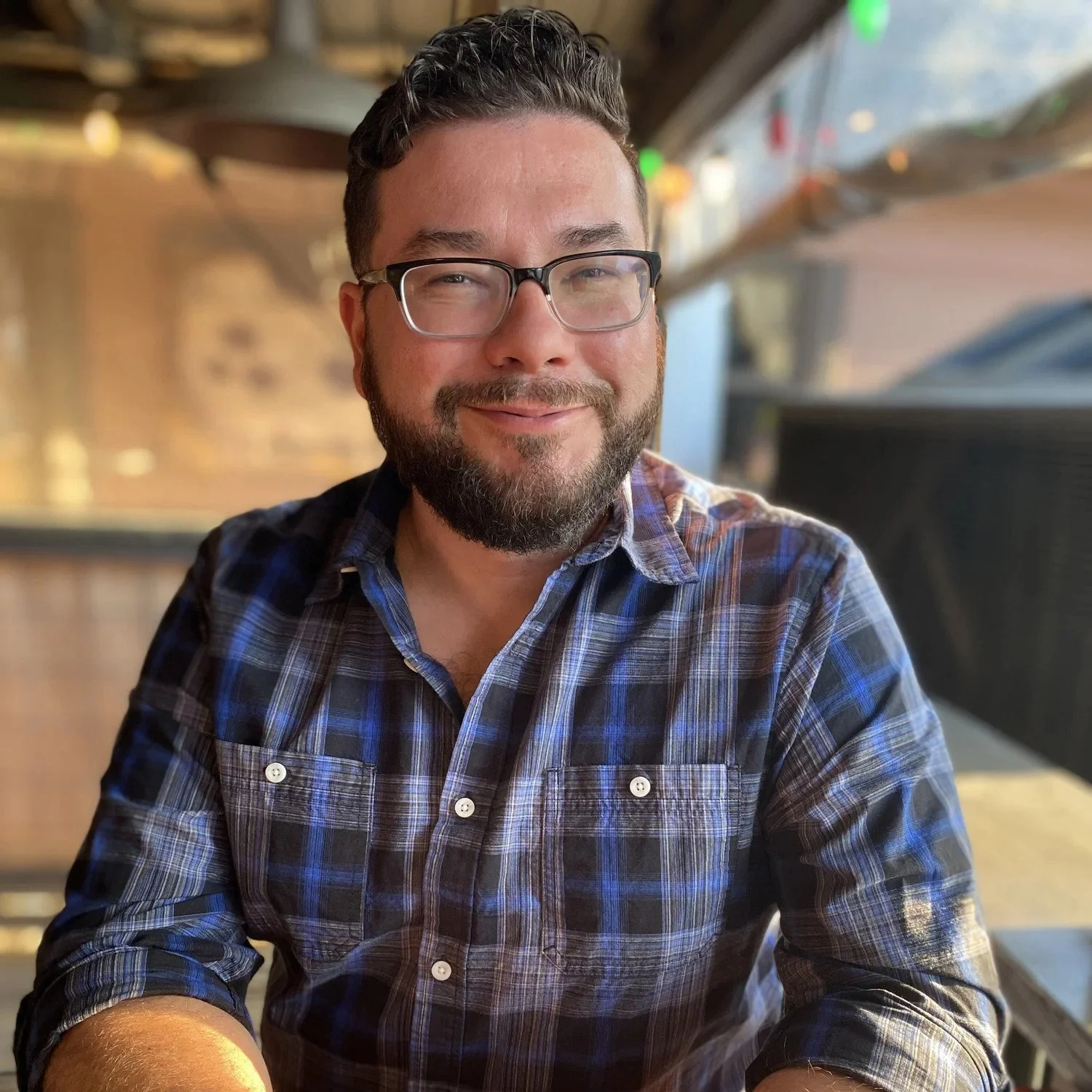 A smiling man with glasses and a beard, wearing a blue plaid shirt, sitting at a table in a restaurant or cafe with a blurred background.