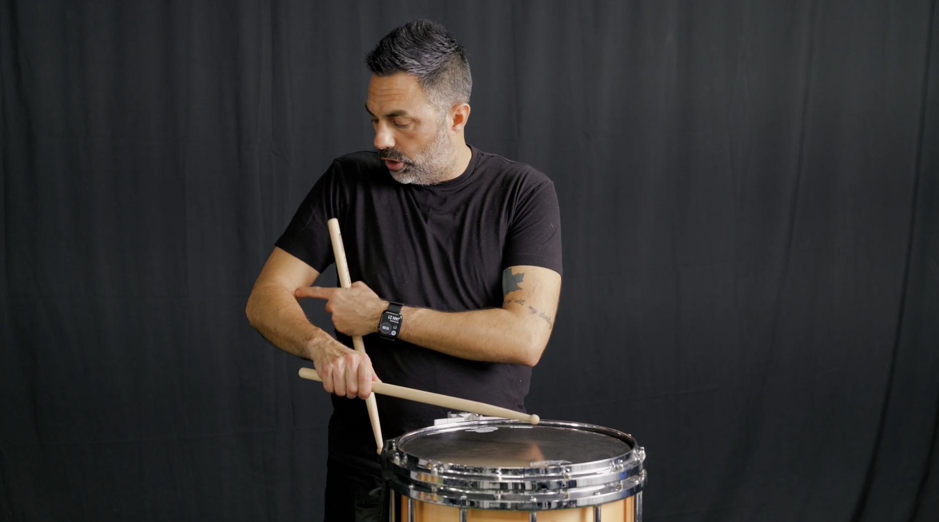 A man performing a stretching exercise indoors on a green turf surface with a black curtain background.