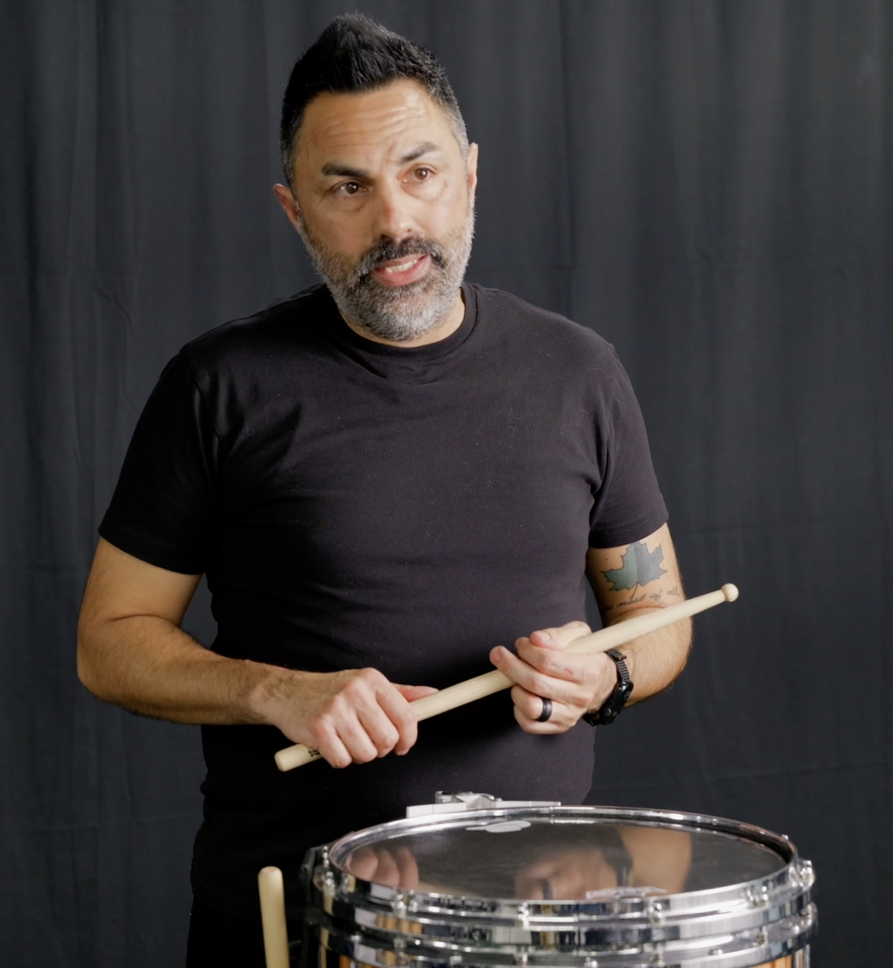 A man with a beard and tattoos holding drumsticks next to a snare drum, speaking in front of a dark backdrop.