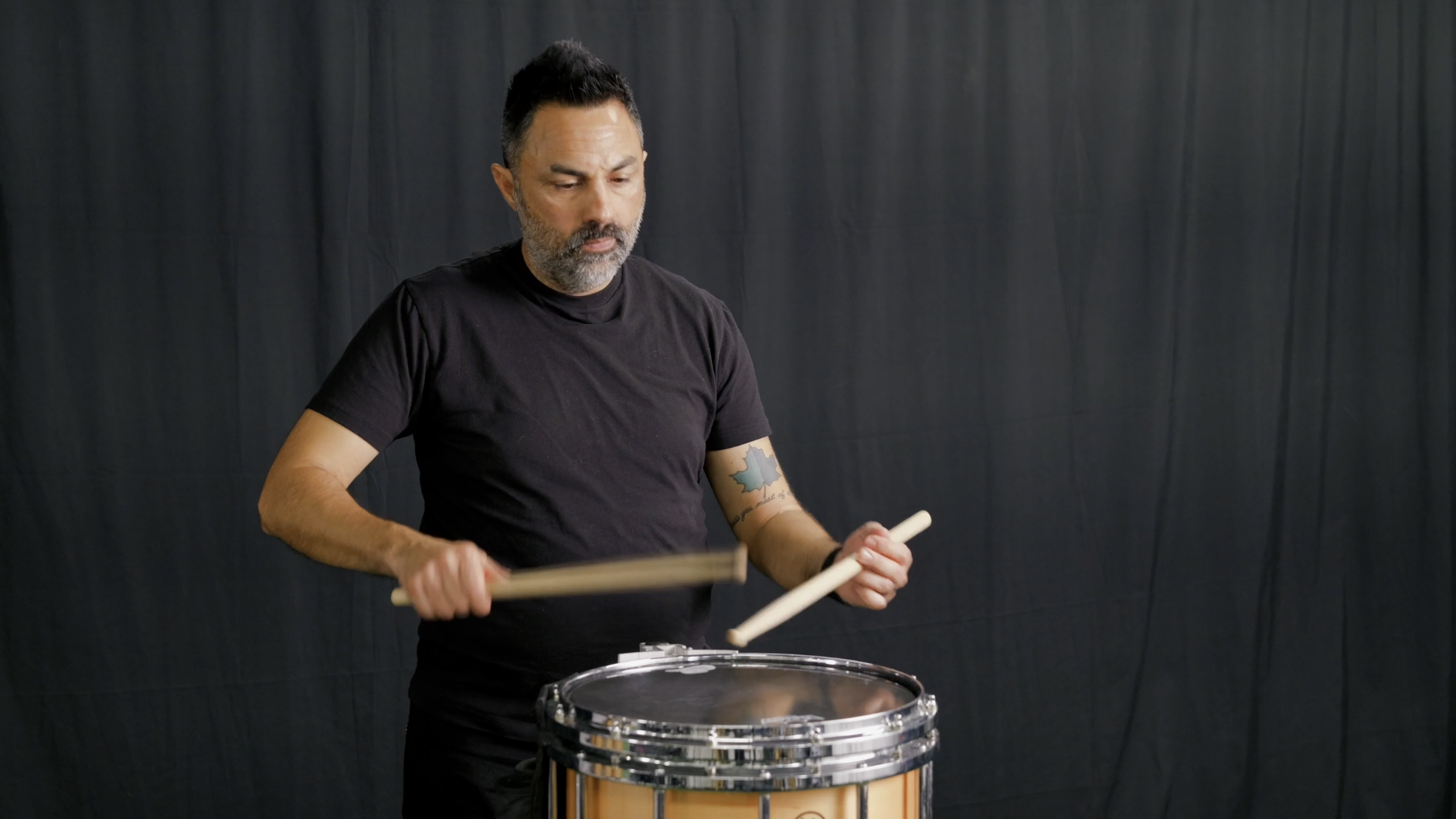 Man squatting on green carpet against black curtain background, wearing black t-shirt, dark pants, and white sneakers.