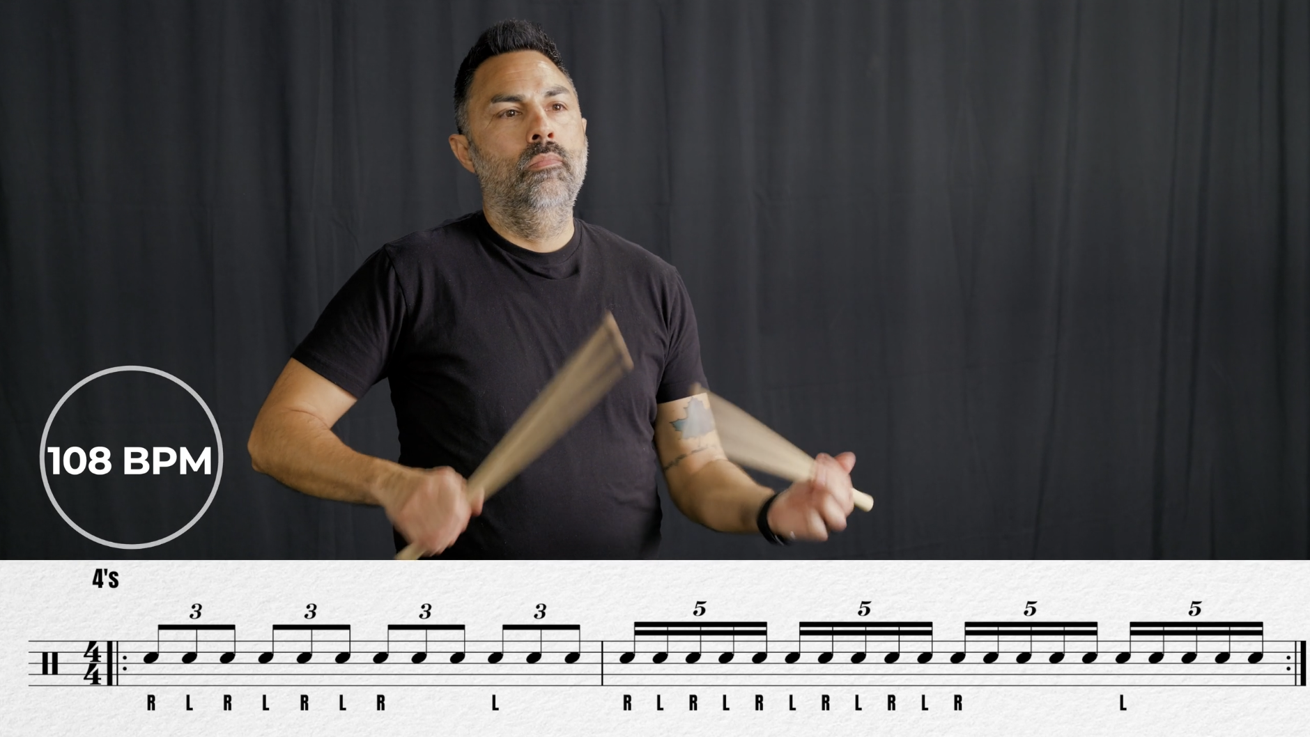 Man standing on green carpet against black curtain, pointing downward with his right hand, wearing black T-shirt, black shorts, black athletic shoes, and a backwards black cap.