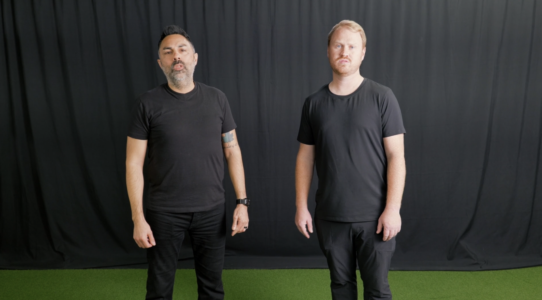 Two men standing on a green artificial turf in front of a black curtain, both wearing black T-shirts and shorts or pants, one with white sneakers and the other with black sneakers, facing the camera.