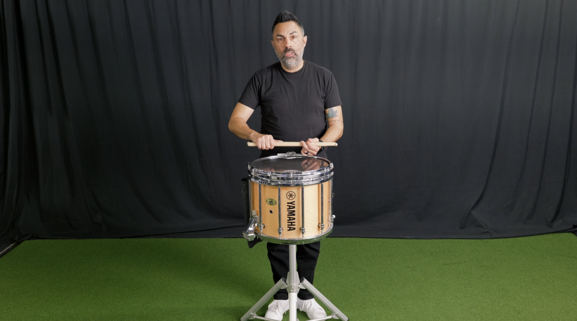 A young man in a black t-shirt and backwards baseball cap standing against a black background, gesturing with his hands as if explaining something.