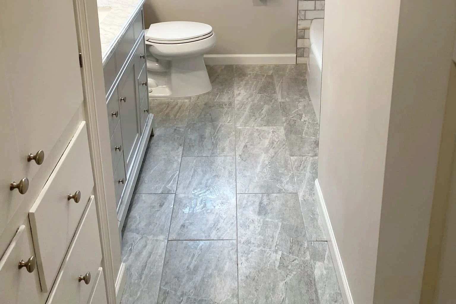 A bathroom with gray tiled floor, white and gray cabinets, a white toilet, and part of a bathtub visible.