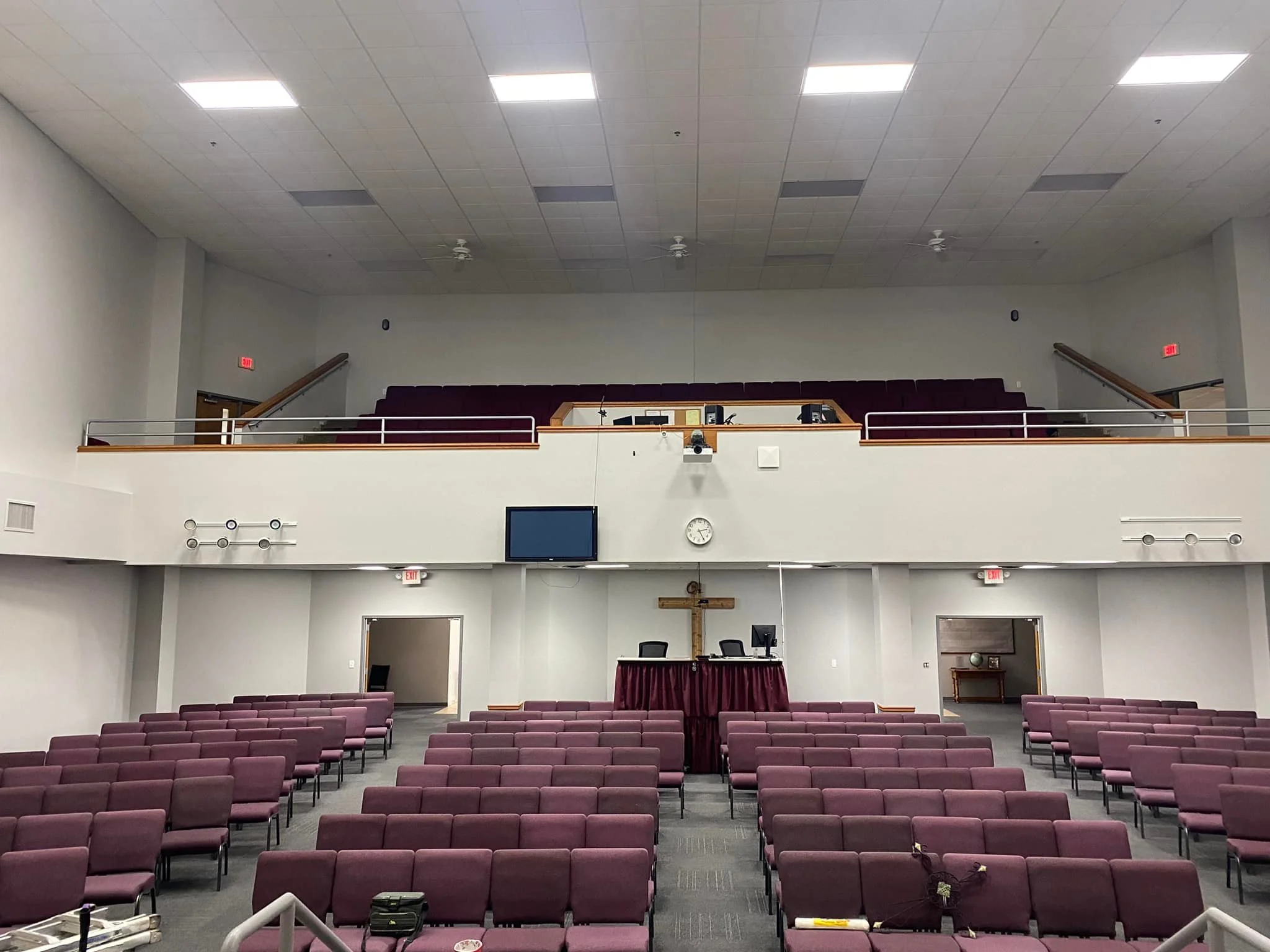 Interior of a church or auditorium with rows of purple chairs, a stage with two chairs and desks behind a purple skirt, a large wooden cross on the wall, and a balcony with more seating. The room is well-lit with ceiling lights and has a clock on the wall.