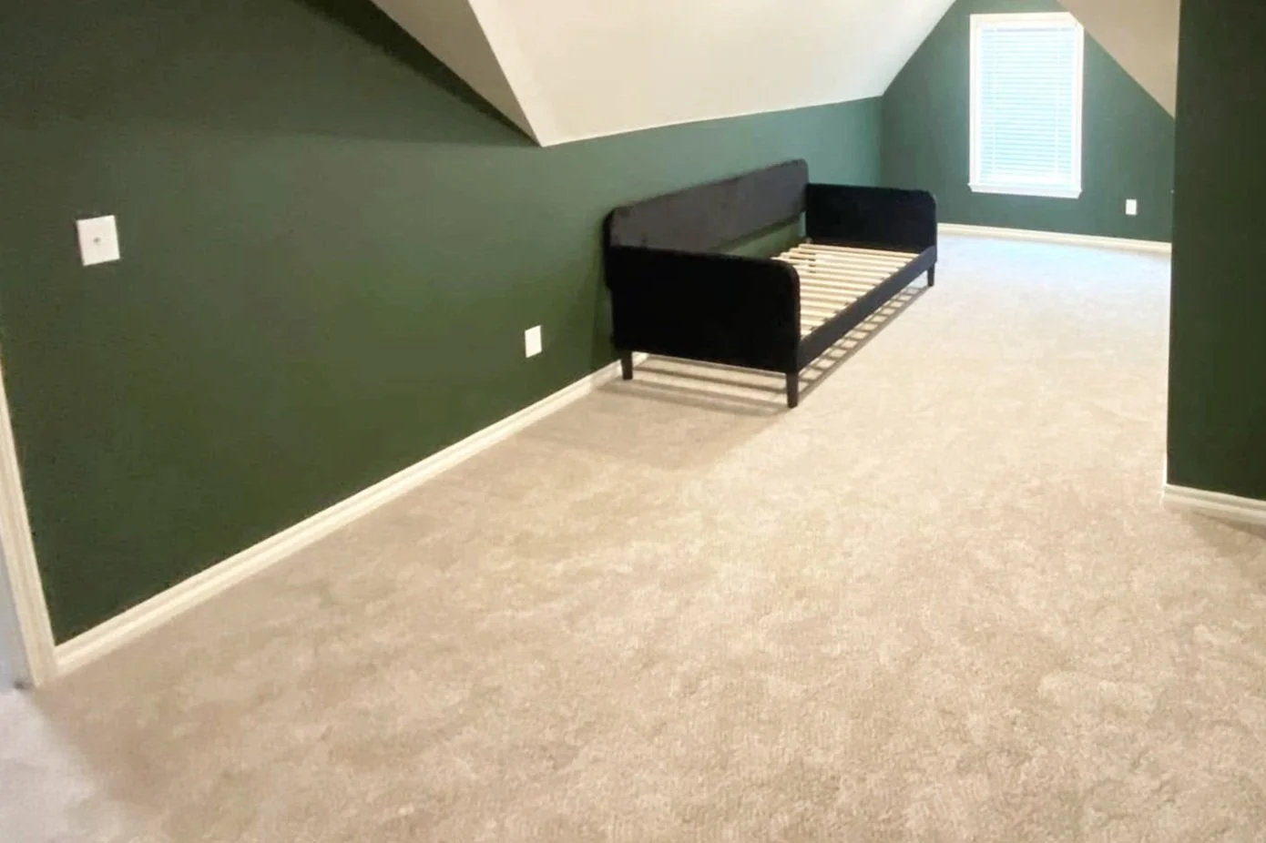 Empty attic room with green walls, a black bed frame without a mattress, beige carpet, a window with blinds, and electrical outlets on the wall.