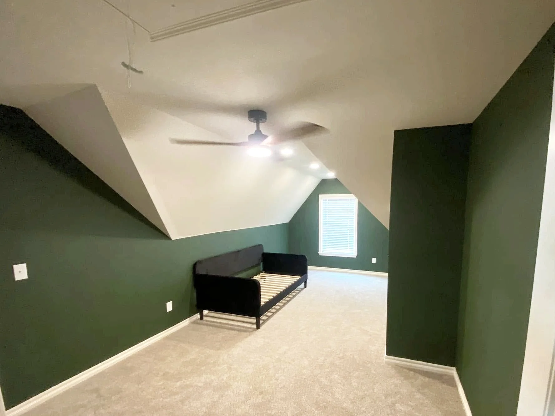 Empty room with green walls, beige carpet, a window with blinds, ceiling fan, and a black bed frame.