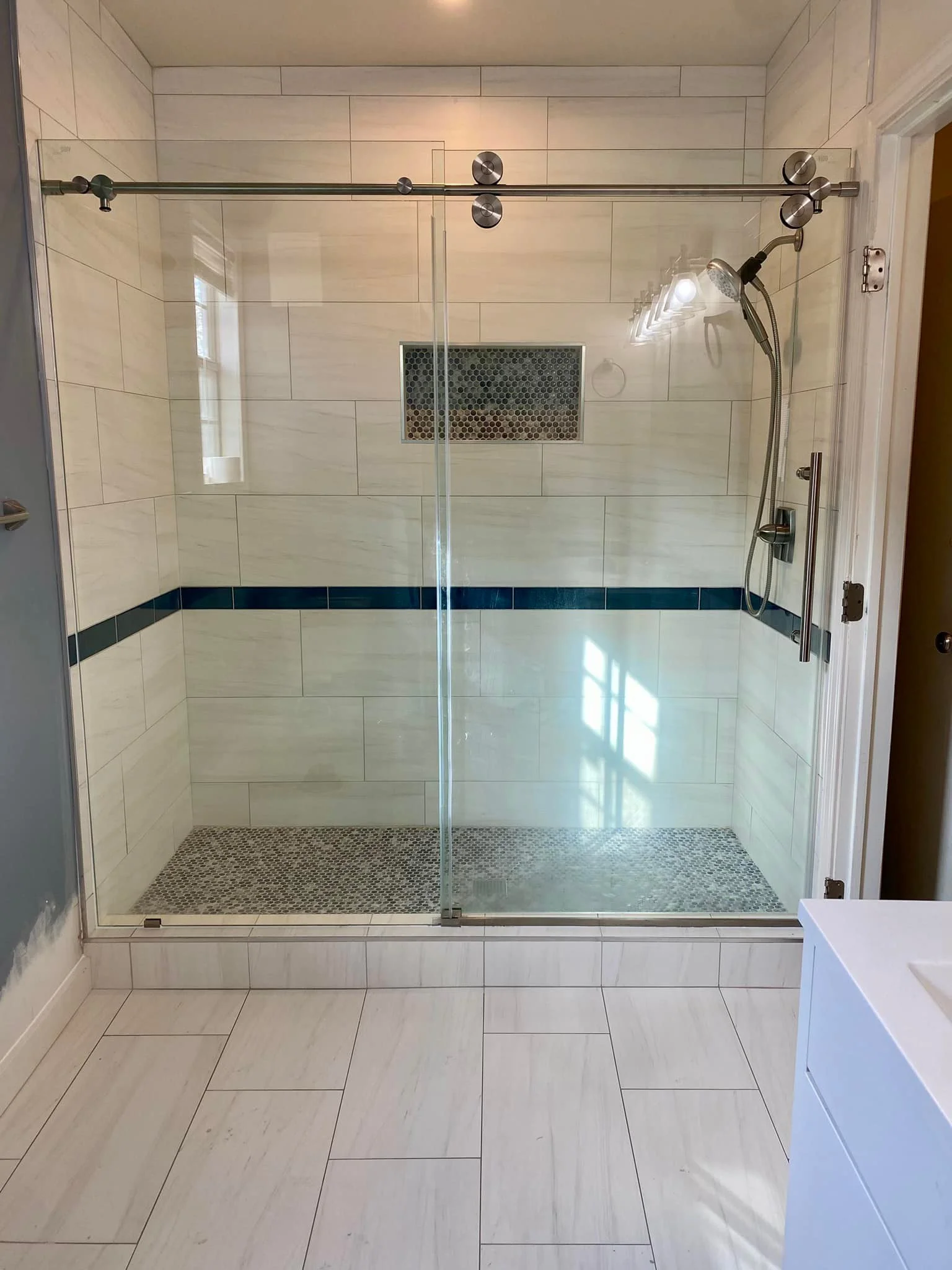 A glass-enclosed walk-in shower with a sliding door, featuring white tiled walls with a blue horizontal accent stripe, a built-in niche with decorative tiles, a handheld showerhead, and a rain showerhead. The floor has small pebble tiles.