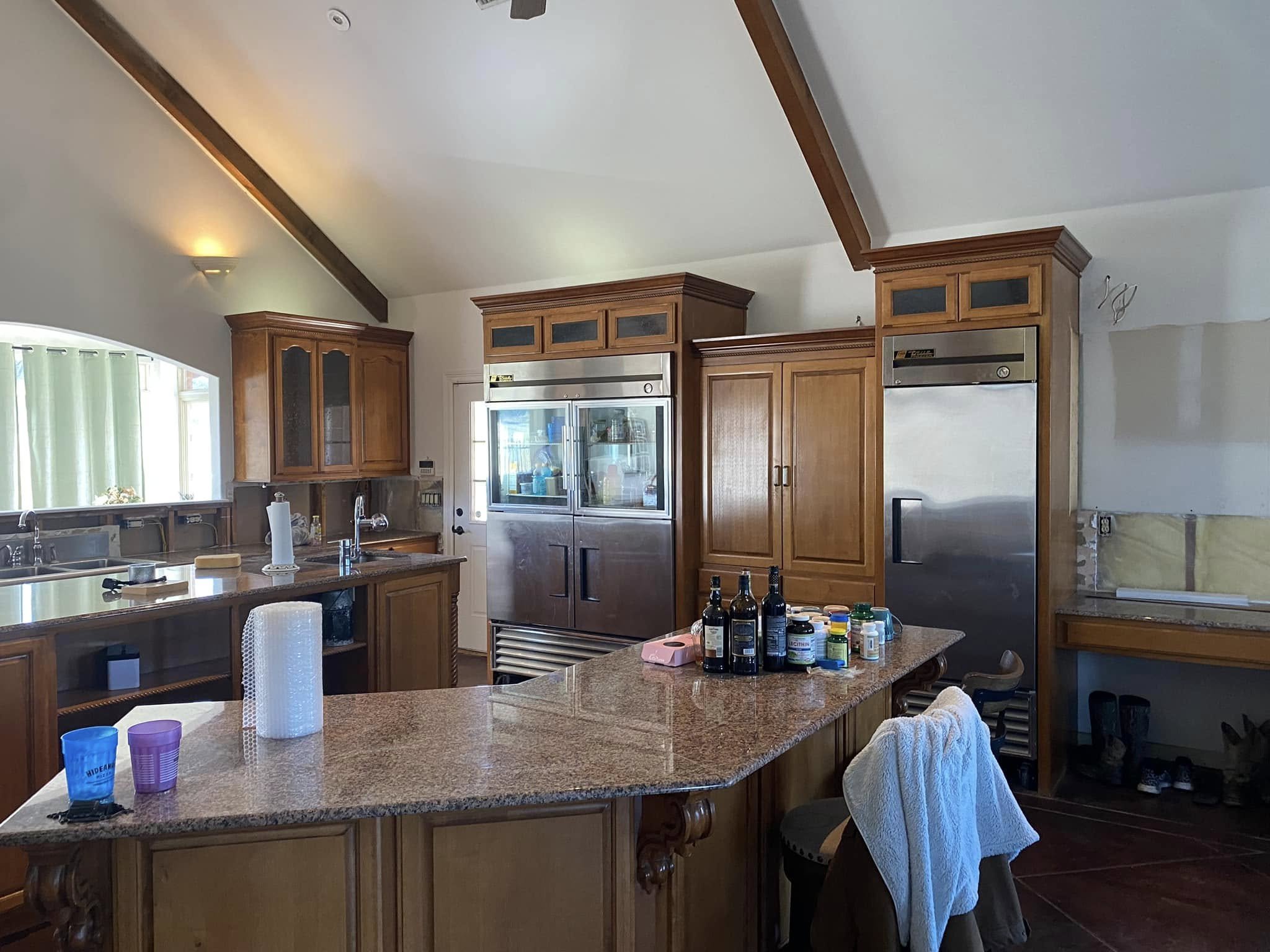 Kitchen with wooden cabinets, stainless steel refrigerator, granite countertop island, and various bottles and pills on the counter. There are paper towels, cups, and a towel on the chair.