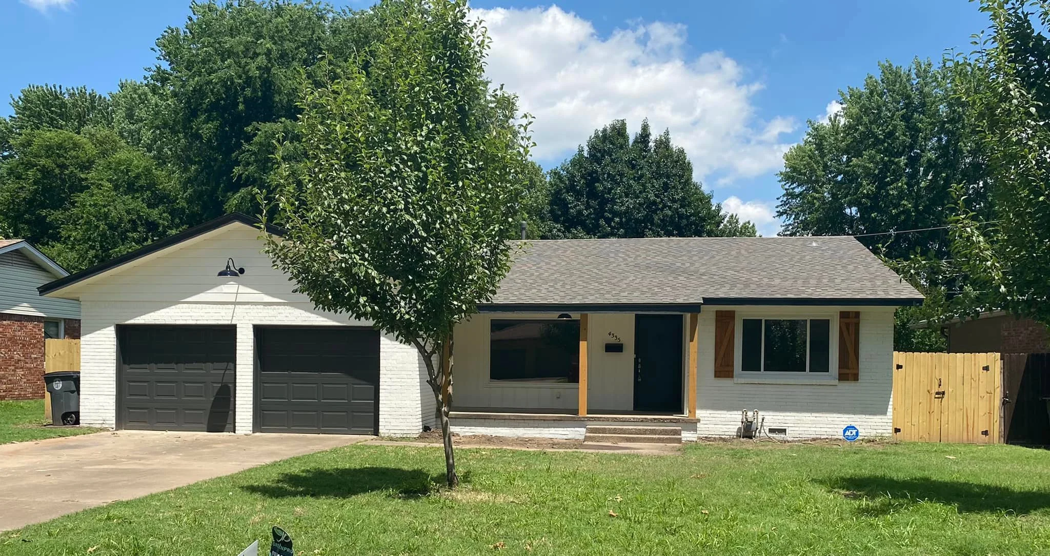 Front view of a single-story house with white brick exterior, black garage doors, and a small front porch with stairs, surrounded by green grass and trees, under a partly cloudy blue sky.