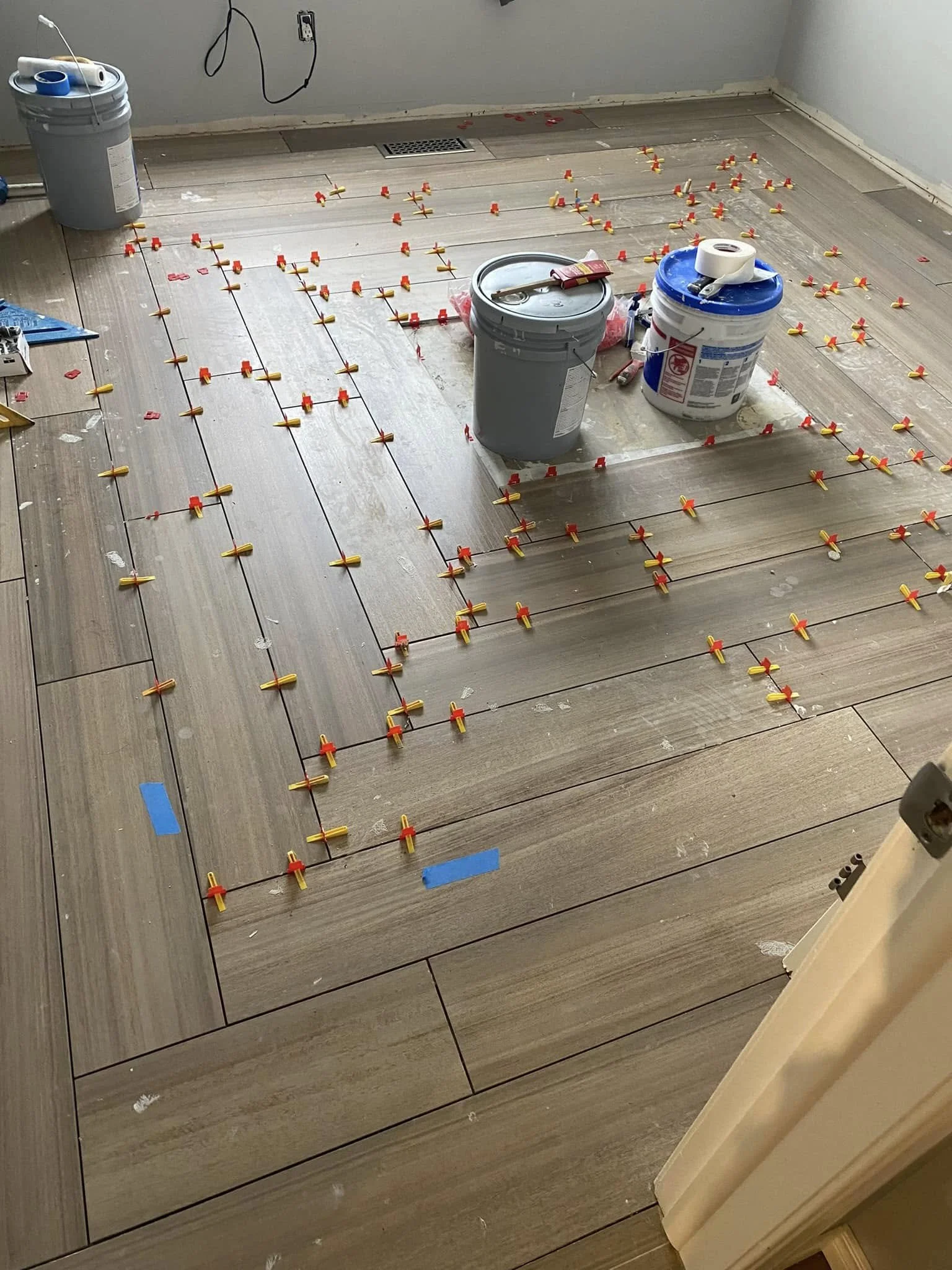 A room with hardwood flooring under renovation, with tile spacers and marked areas for tiling installation, various buckets and tools for tile work, a floor vent, and a small pile of construction materials.