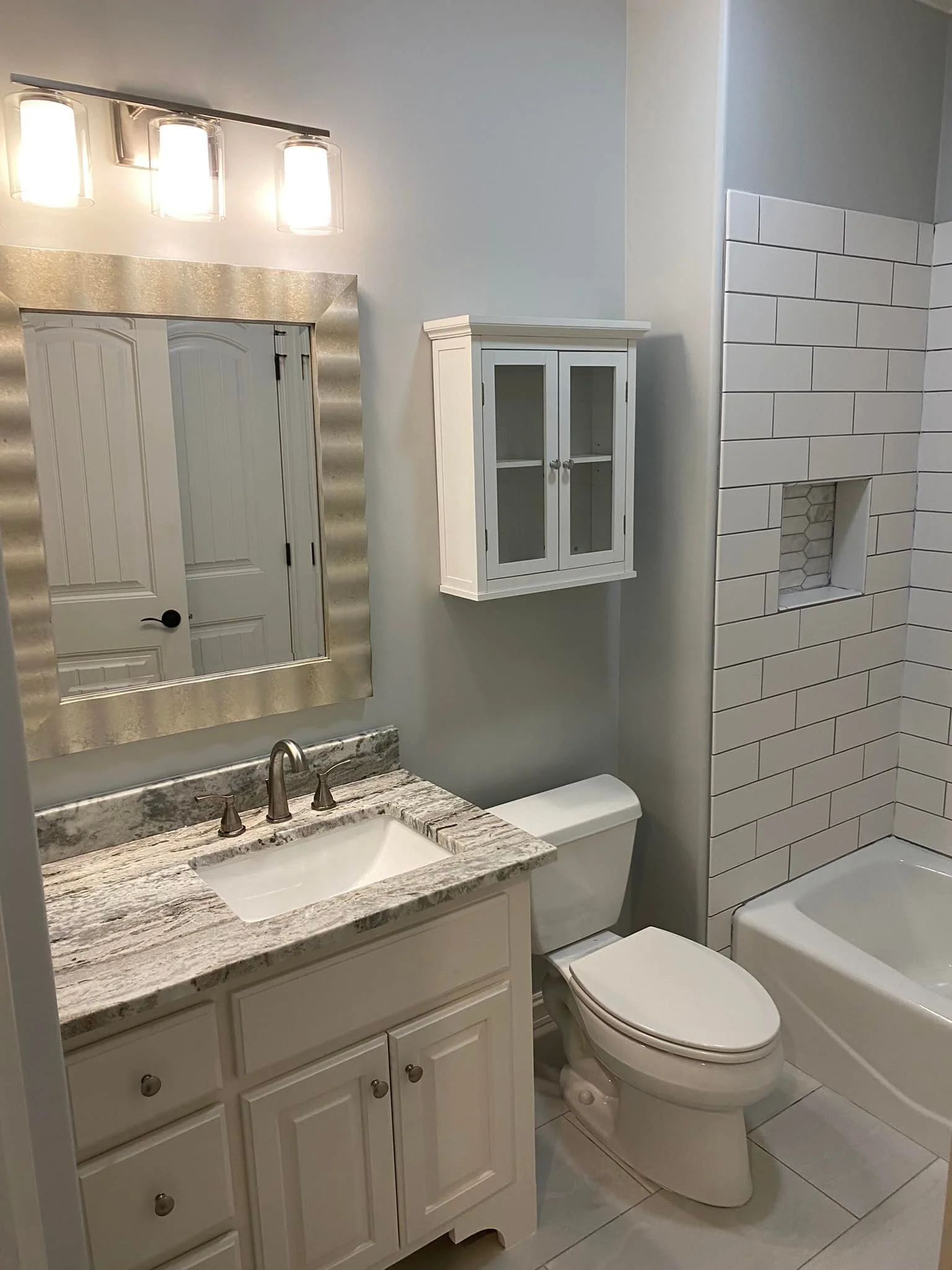 A bathroom with a granite countertop vanity, a mirror with a silver frame, a white toilet, a wall-mounted white glass cabinet, and a bathtub with white tile surround and built-in niche.