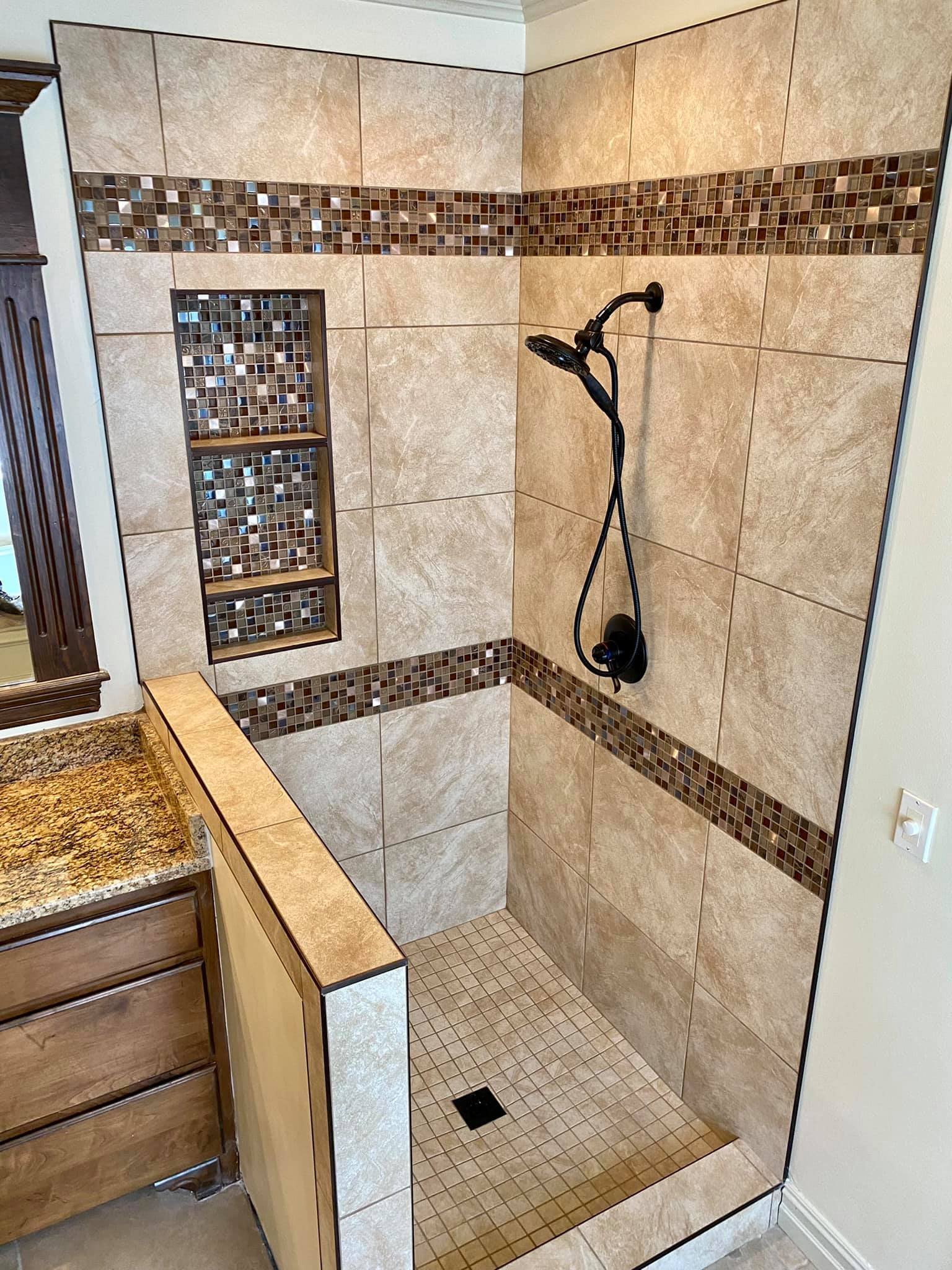 A walk-in shower with beige tiles, a mosaic tile border, a built-in niche with shelves, a black handheld showerhead, and a drain in the floor.