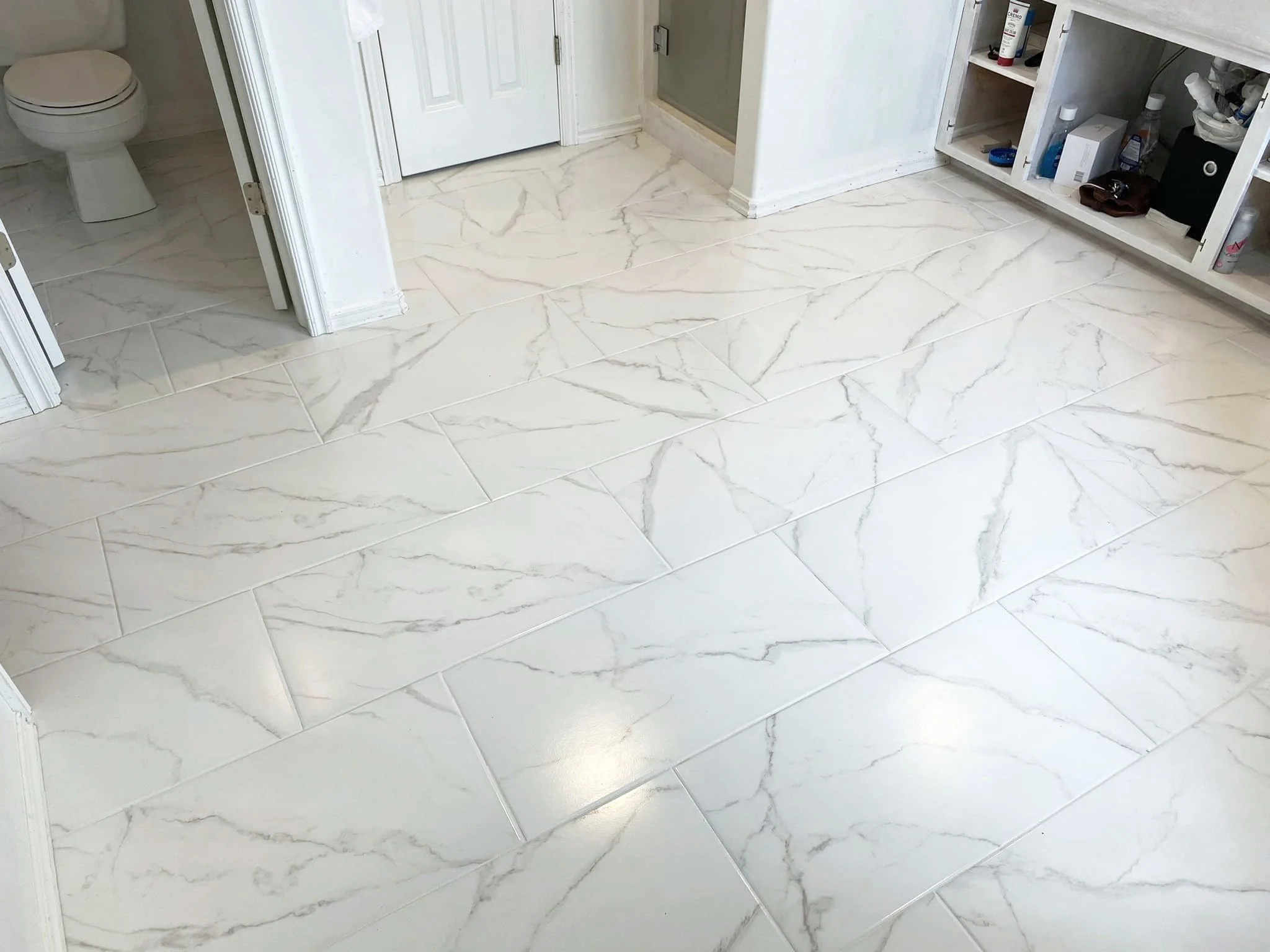 Bathroom with white marble tile flooring, a toilet in the corner, a partially open door, and a white cabinet with bathroom supplies.