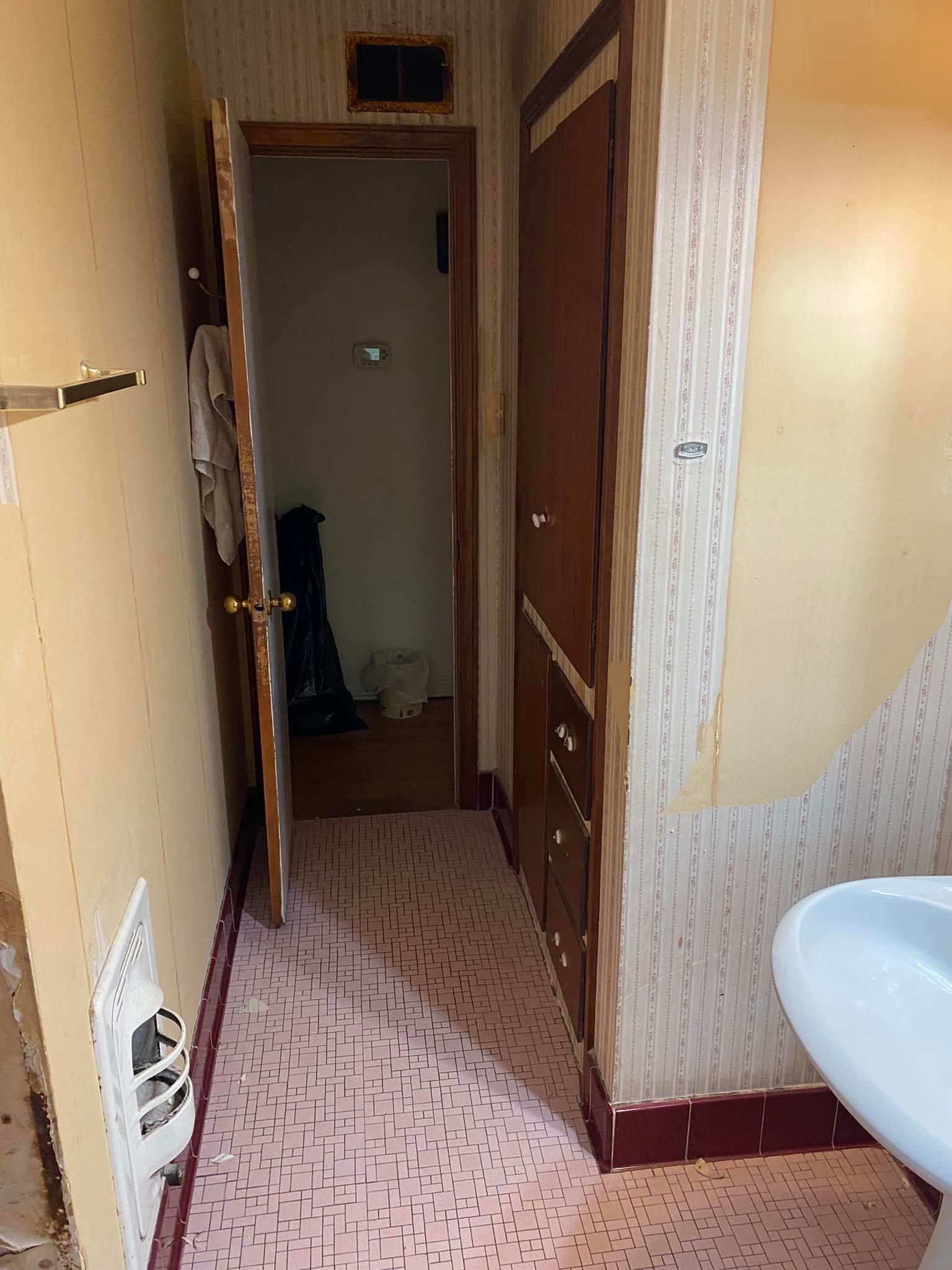 A small bathroom with pink tile flooring, partial yellow walls, a white sink, wooden cabinets, a doorway leading to a narrow hallway, a towel hanging on a hook, and a waste basket in the hallway.