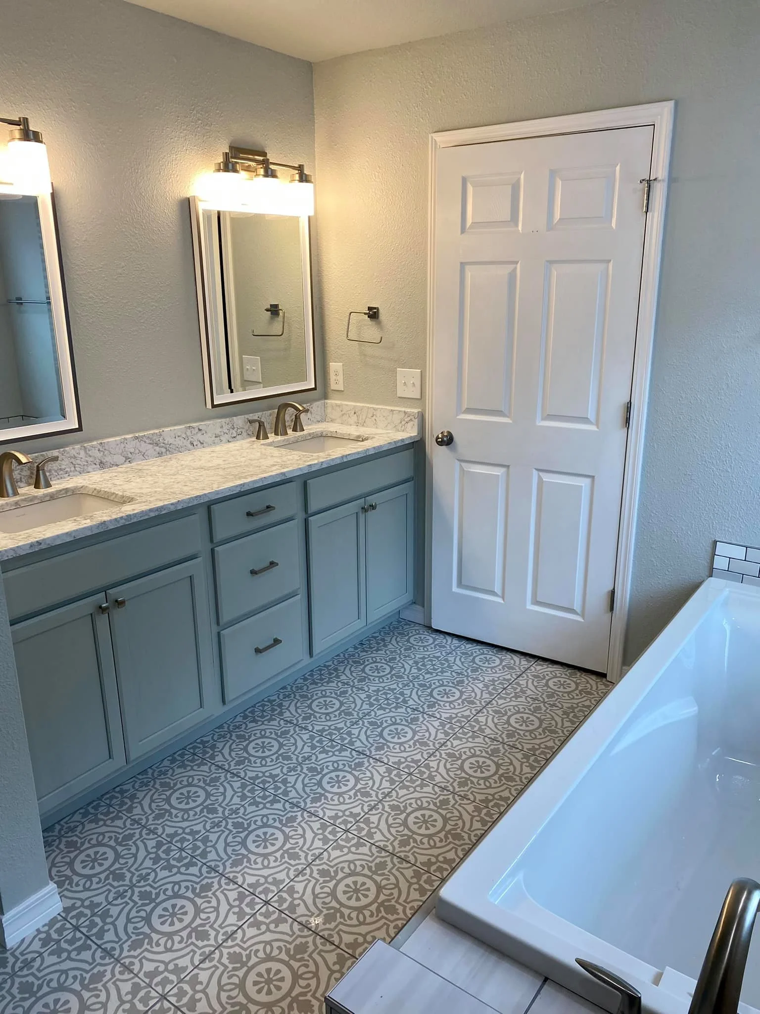 A modern bathroom featuring a double vanity with light gray cabinets, white marble countertop, and two oval sinks. There are two mirrors above the sinks with light fixtures, and a closed white door next to a bathtub on the right. The floor has decora