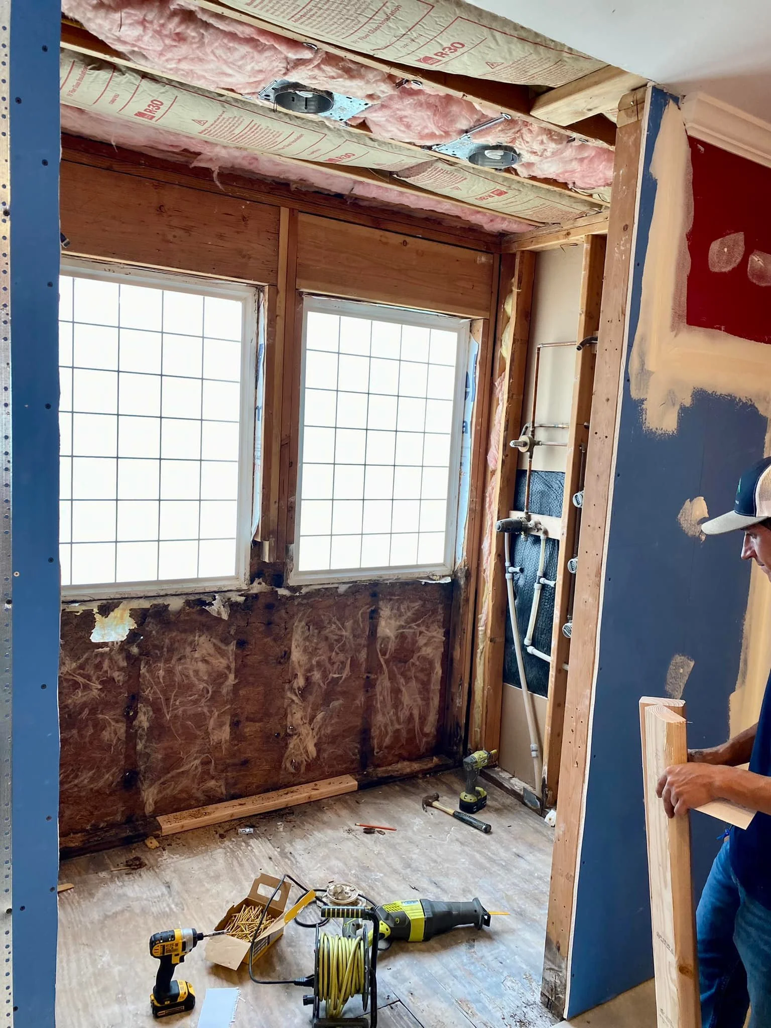 Construction room with two windows, insulation, plumbing pipes, and various tools including a drill, extension cord, and screws. A person is working on a wooden frame.