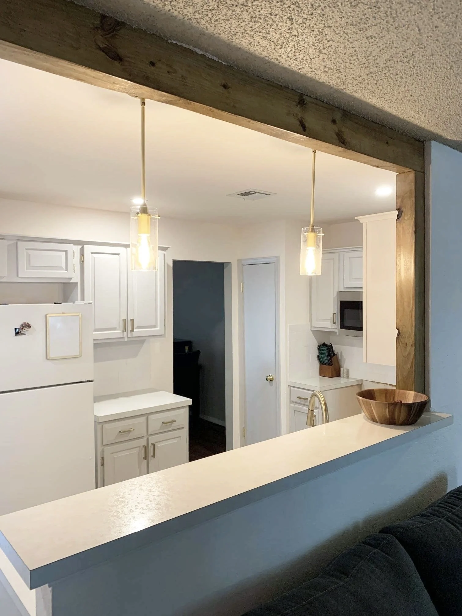 View of a kitchen seen through a bar opening, with white cabinets, a white refrigerator, a microwave, a counter with a wooden bowl, and hanging pendant lights.