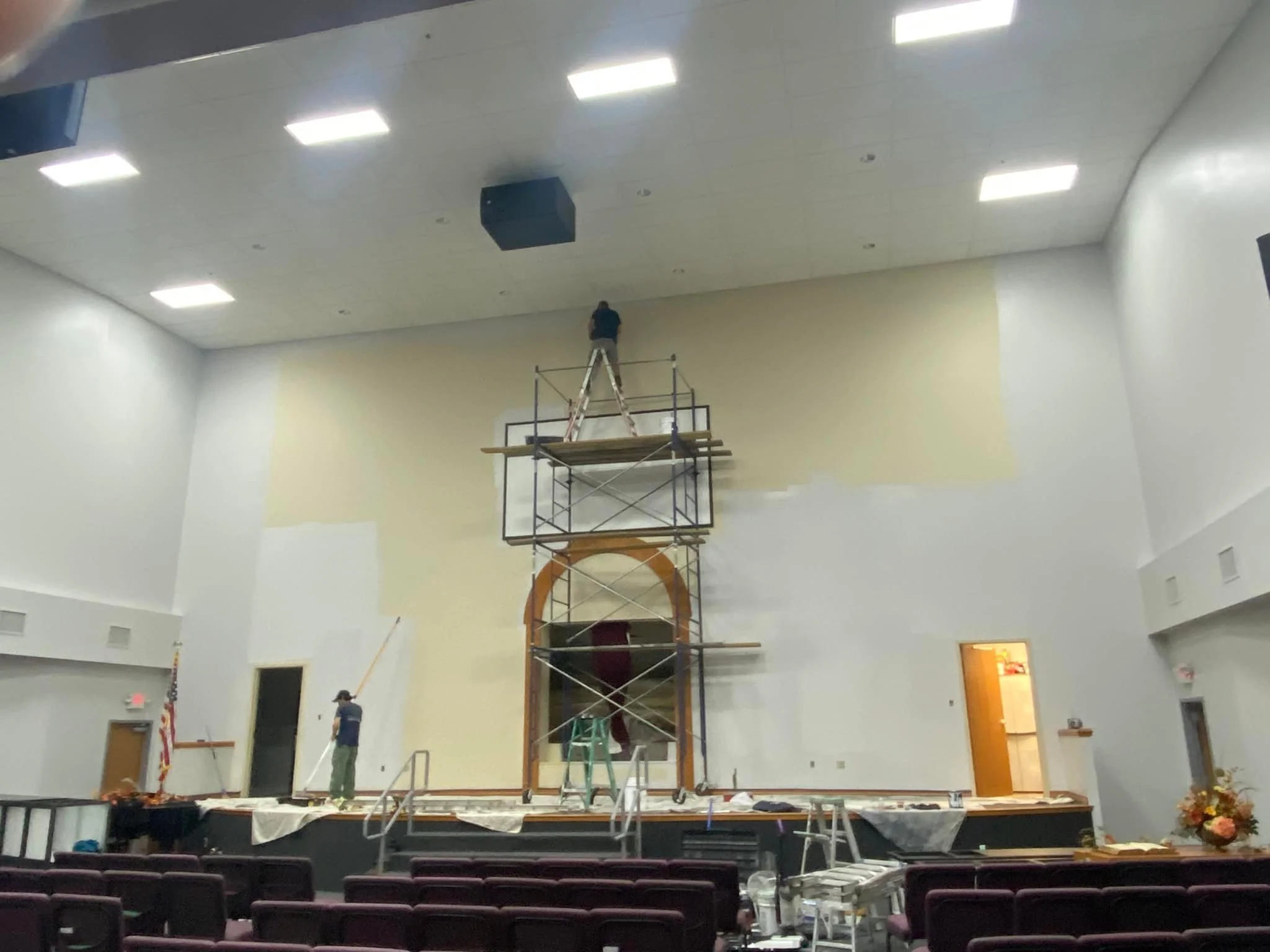 People working on scaffolding in a church or auditorium stage area, painting or repairing the wall. The stage has chairs and a floral arrangement to the side.