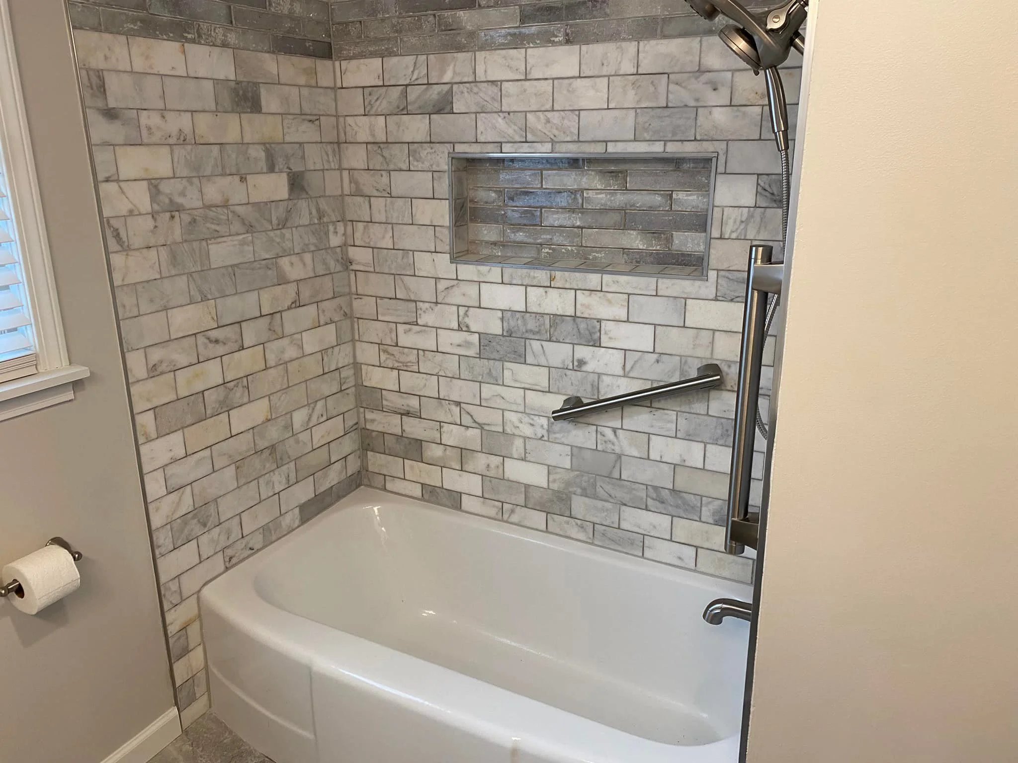 A bathtub in a bathroom with a tiled walk-in shower. The tiles are a mix of white and gray marble. There is a built-in shower shelf and a metal grab bar on the wall.