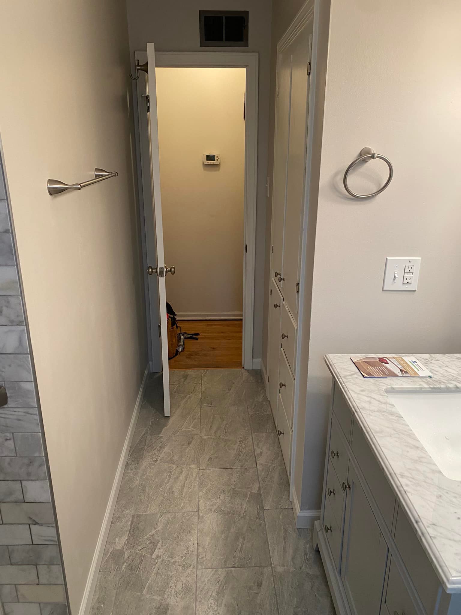 View of a bathroom hallway with a tile floor, white cabinets, a marble countertop with a sink, towel rings on the wall, and an open door leading to an entryway with a thermostat on the wall.